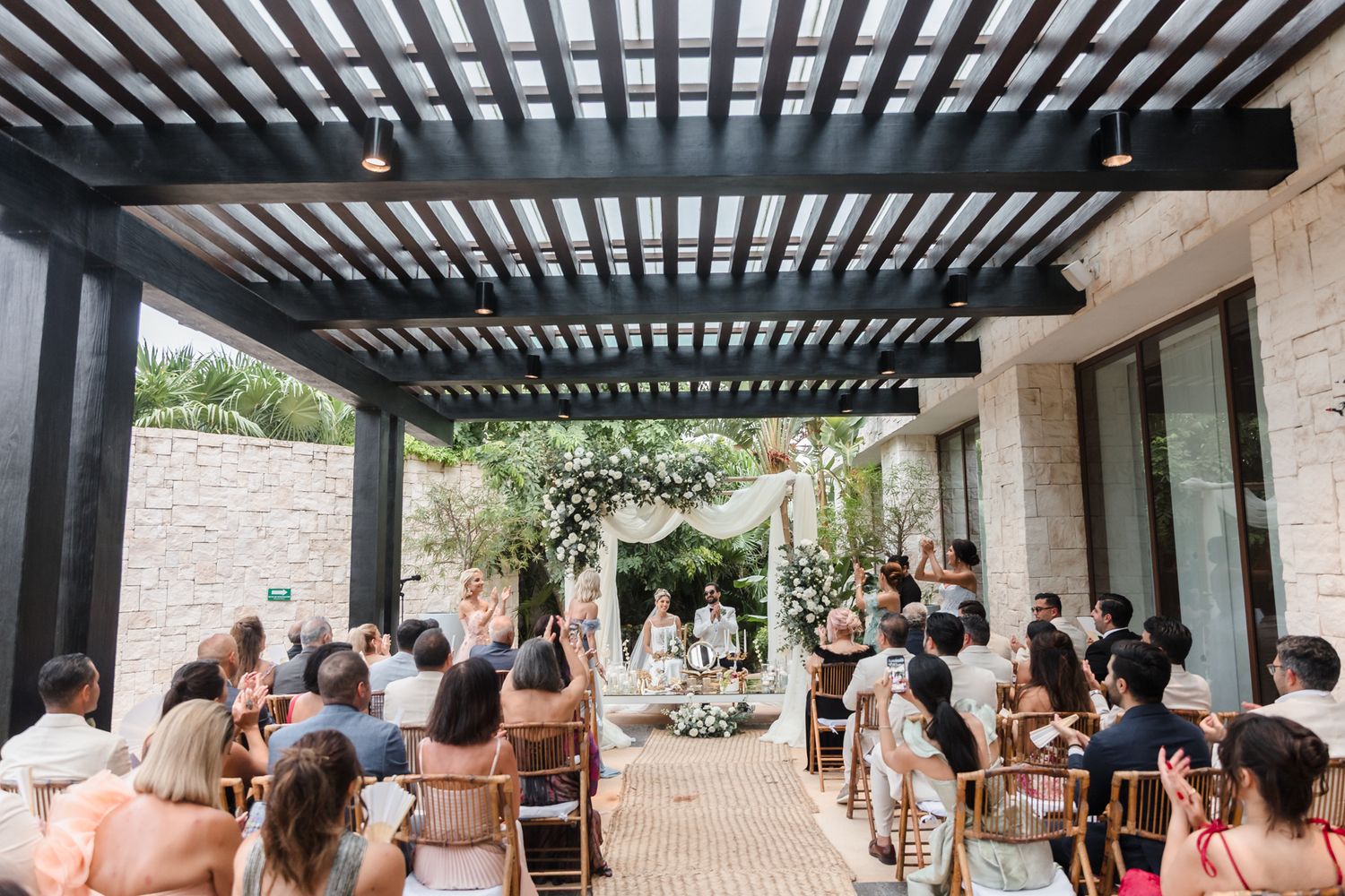 Outdoor wedding ceremony under a dark pergola with guests seated in wooden chairs facing a floral arch altar.