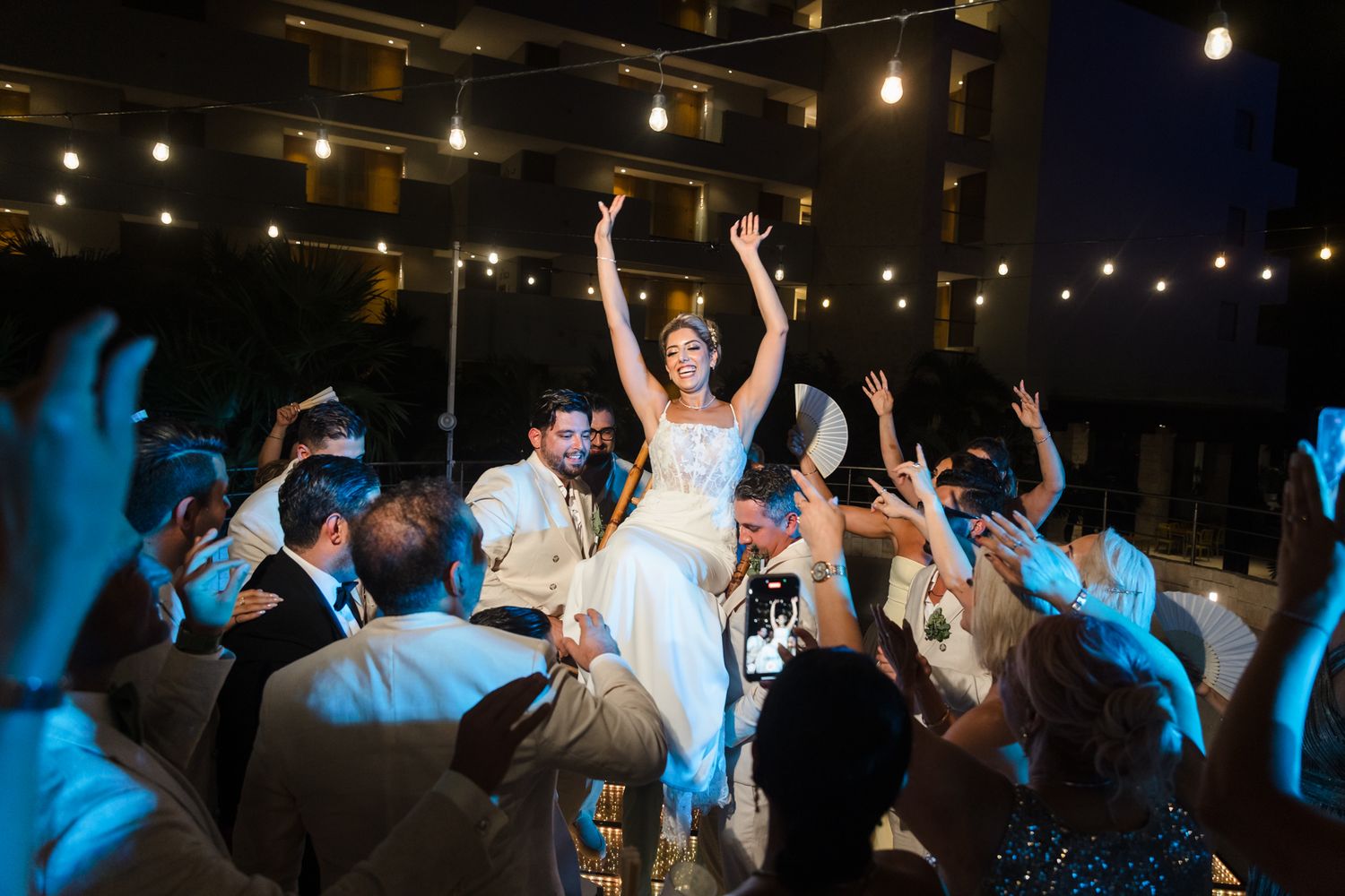 Bride raised in chair during joyful hora dance at outdoor wedding reception at night.