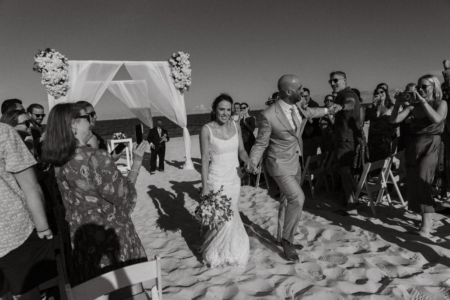 Bride and groom walk down sandy beach aisle after wedding ceremony as guests cheer and photograph.