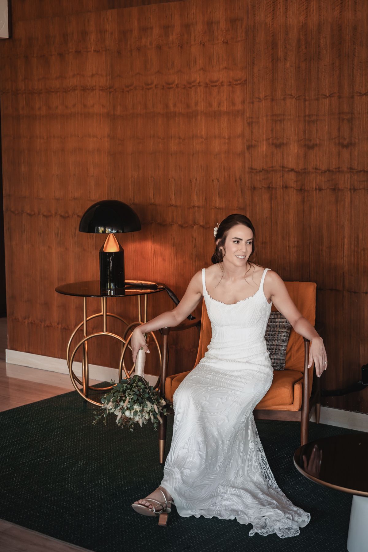 Bride in white lace gown seated in orange chair beside a dark lamp and bouquet against wood-paneled wall.