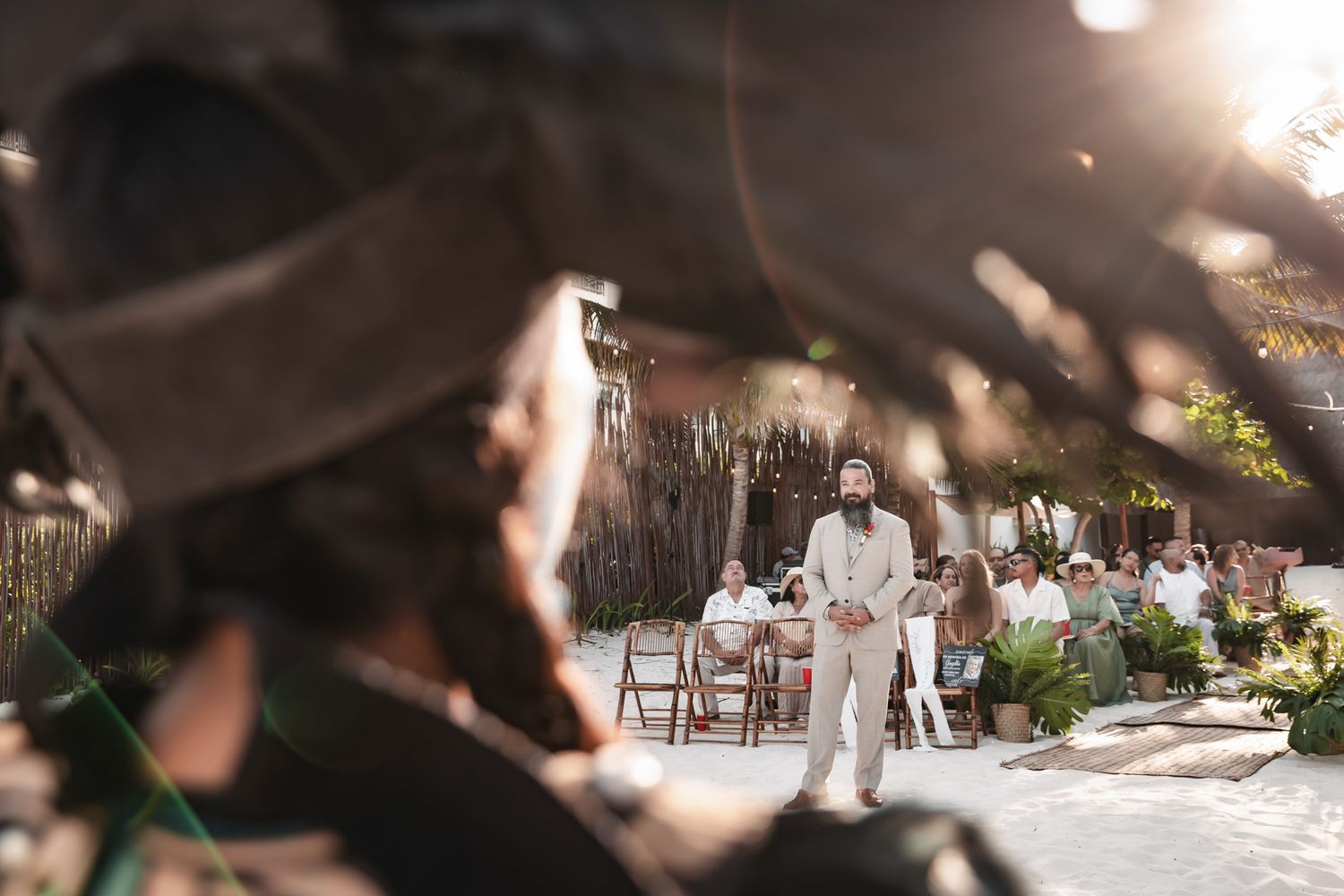 Groom in beige suit stands at outdoor wedding ceremony with guests seated among trees and warm sunlight.