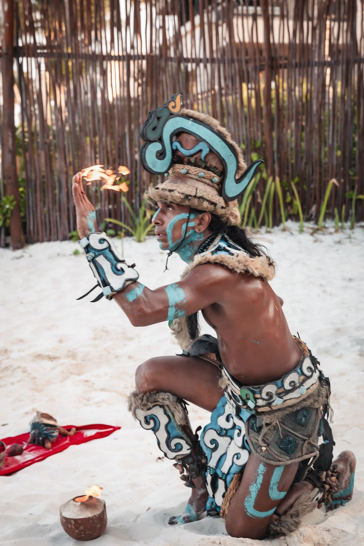 Mayan warrior in traditional blue body paint and headdress performing a fire ritual on a white sand beach.