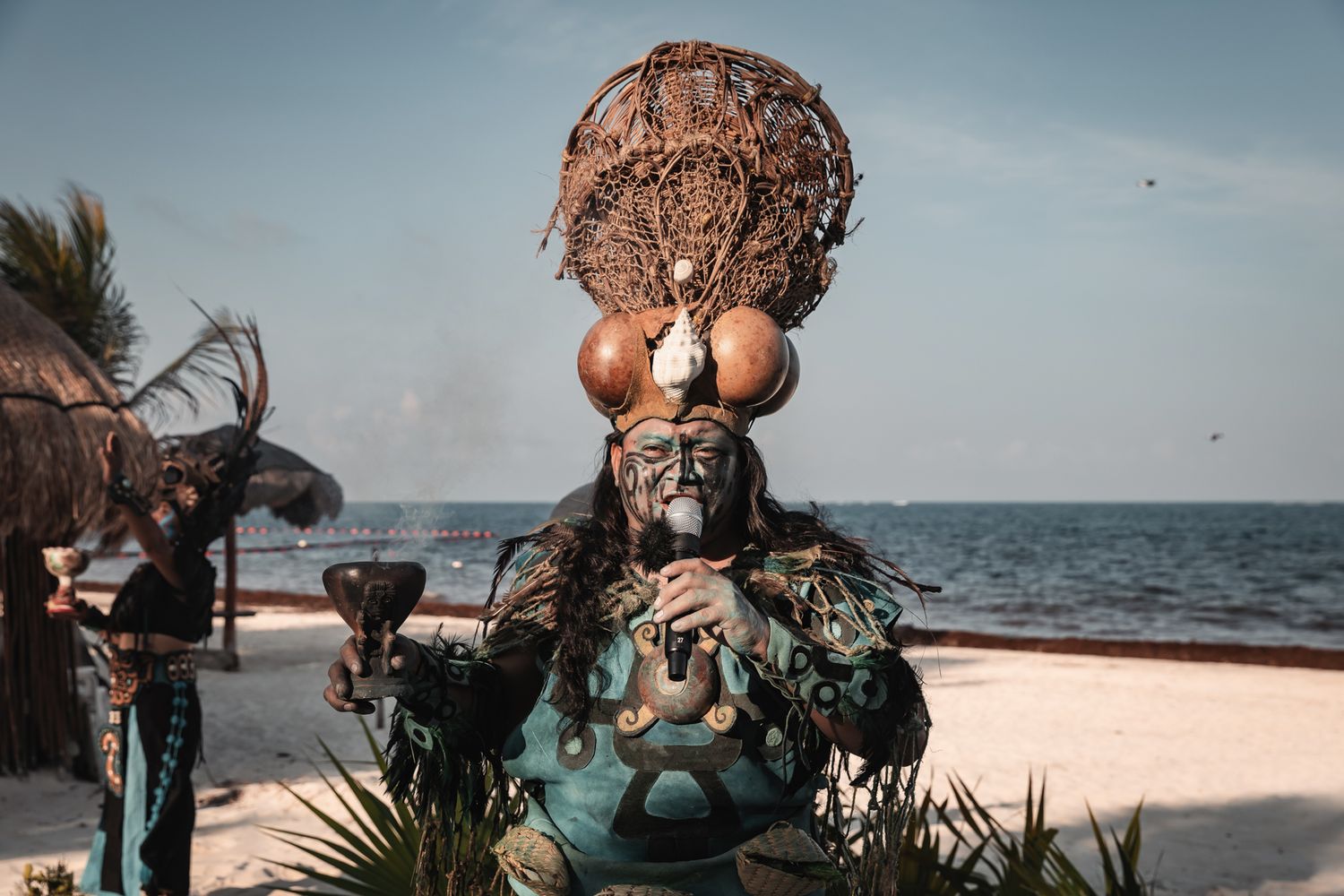 Traditional Mayan shaman in elaborate ceremonial costume on a tropical beach with ocean backdrop.