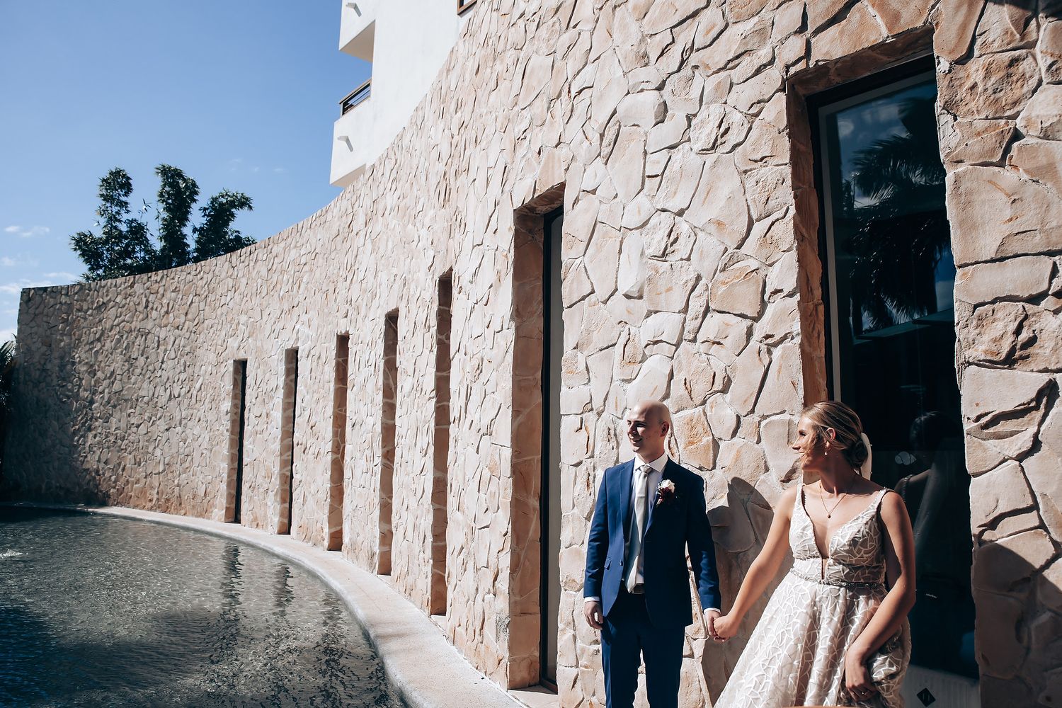 A bride and groom walk together on a stone path, with a stone wall and foliage in the background.