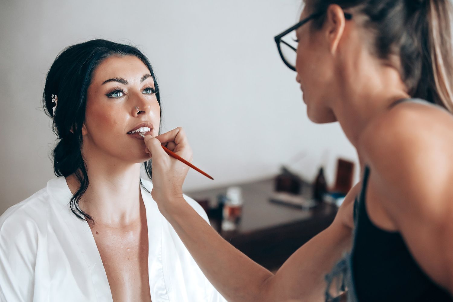 A woman in a white robe is having her makeup applied by another woman in a salon setting.