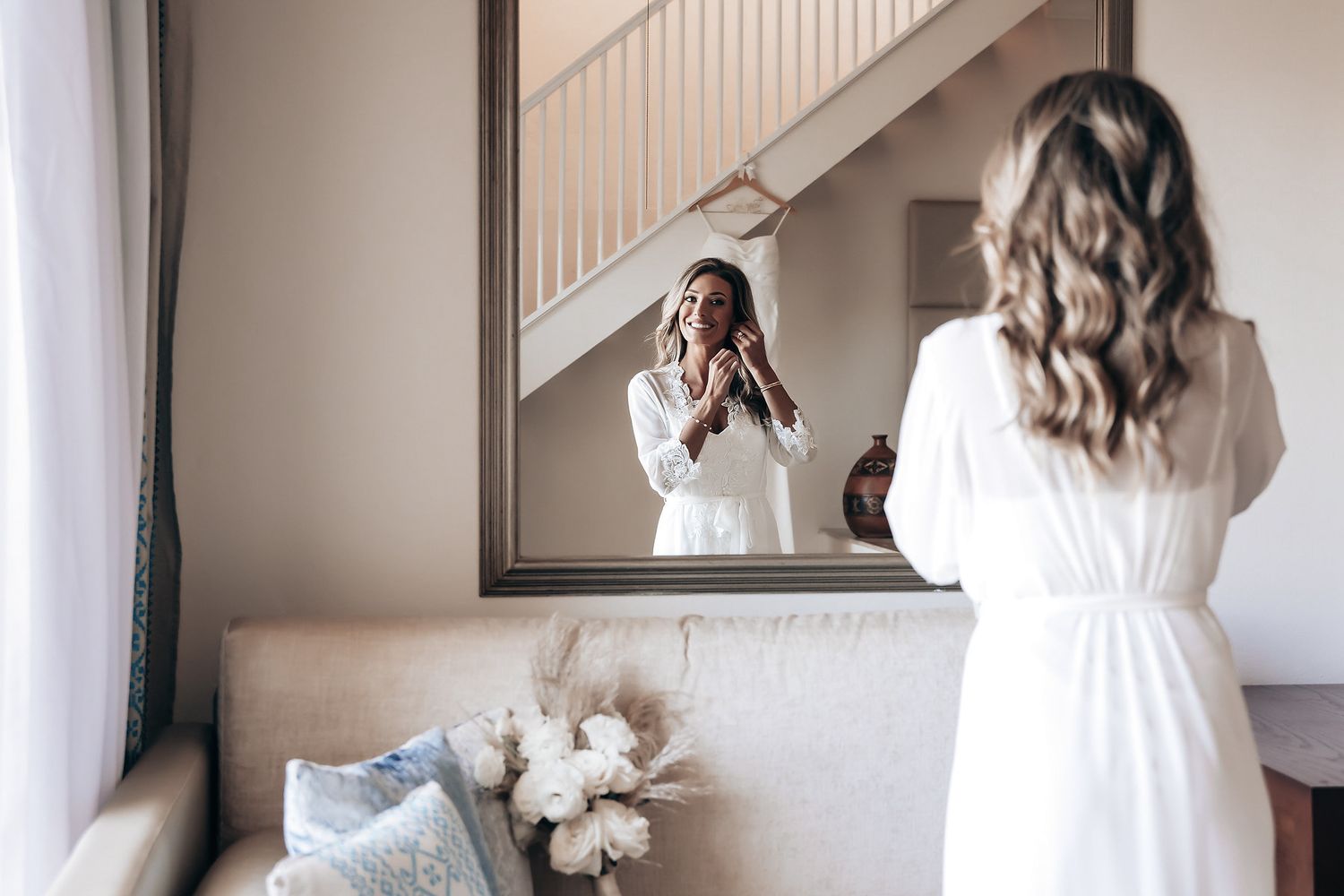 A woman in a white wedding dress is standing in front of a mirror, preparing for her wedding.