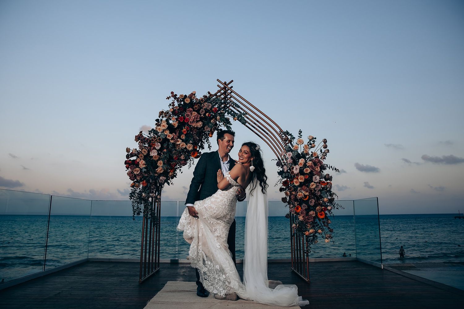 Couple shares romantic moment under floral arch at sunset beach wedding.