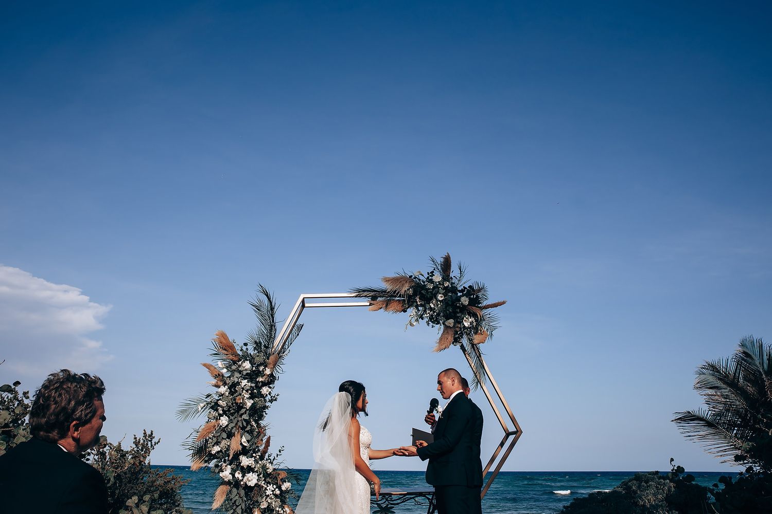 Beachside wedding ceremony under blue skies with wooden arch decorated in florals.
