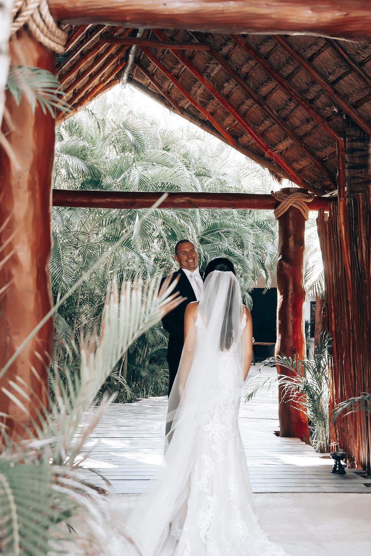 A flowing white wedding dress swirls gracefully under a rustic wooden covered walkway surrounded by palm trees.