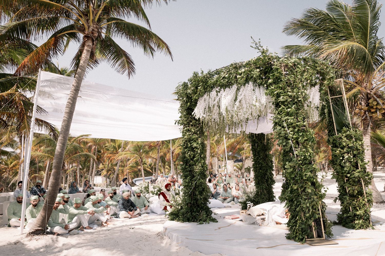 Lush green floral arch on white sand beach wedding ceremony with palm trees and tropical decor.