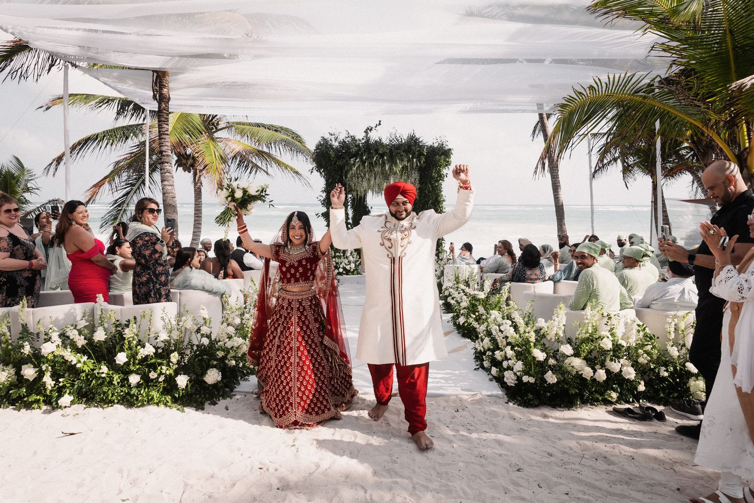 Indian couple in traditional attire celebrating beach wedding ceremony exit with guests.