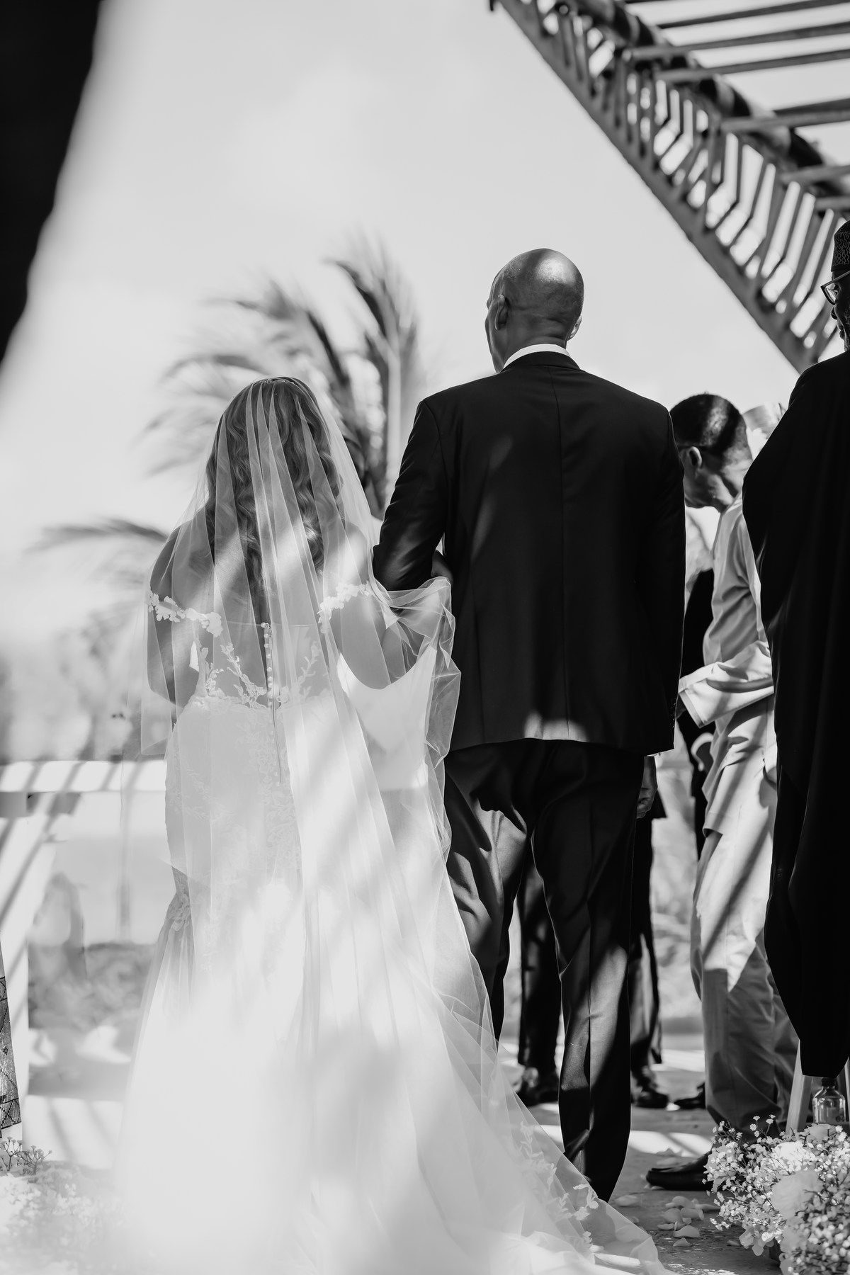 Black and white photo of bride in veil and gown walking down aisle with escort at outdoor wedding.