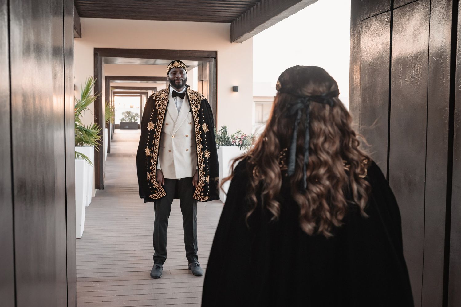 Groom in ornate black and gold ceremonial robe stands in a hallway facing his bride.