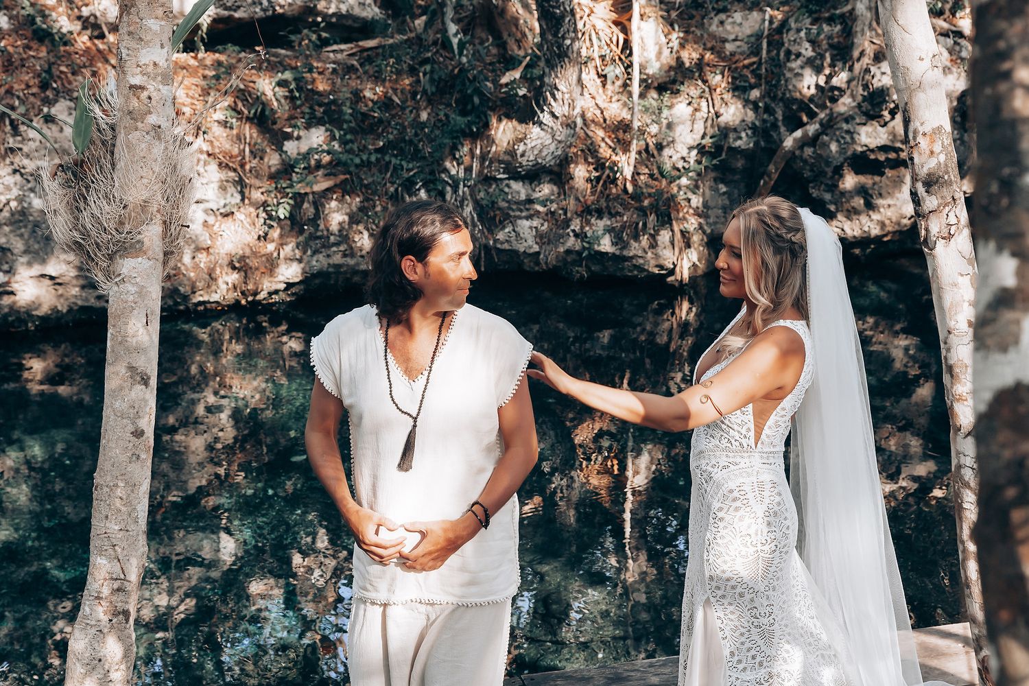 A couple in white outfits poses together in a wooded forest setting during a romantic photoshoot.