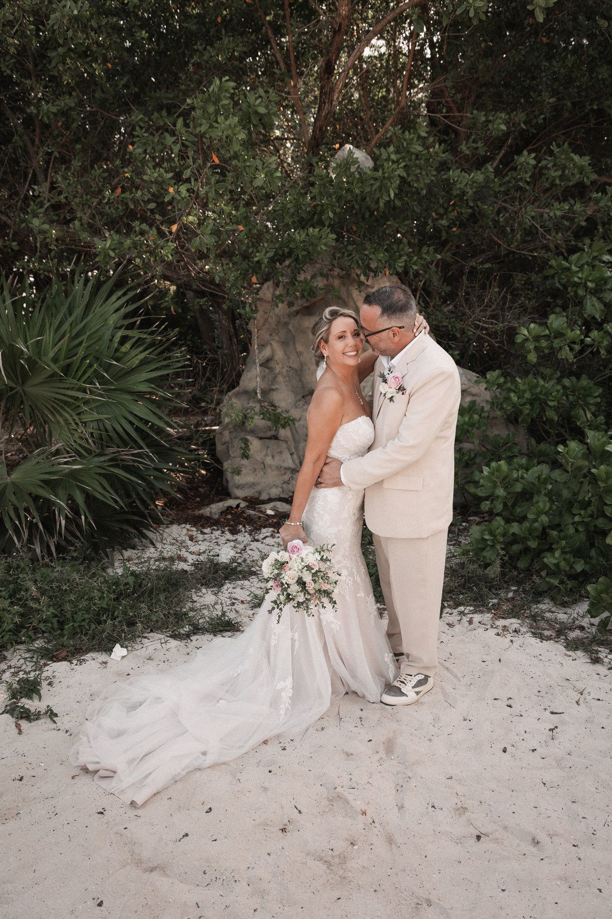 Bride and groom embrace on sandy beach surrounded by lush tropical greenery during outdoor wedding ceremony.
