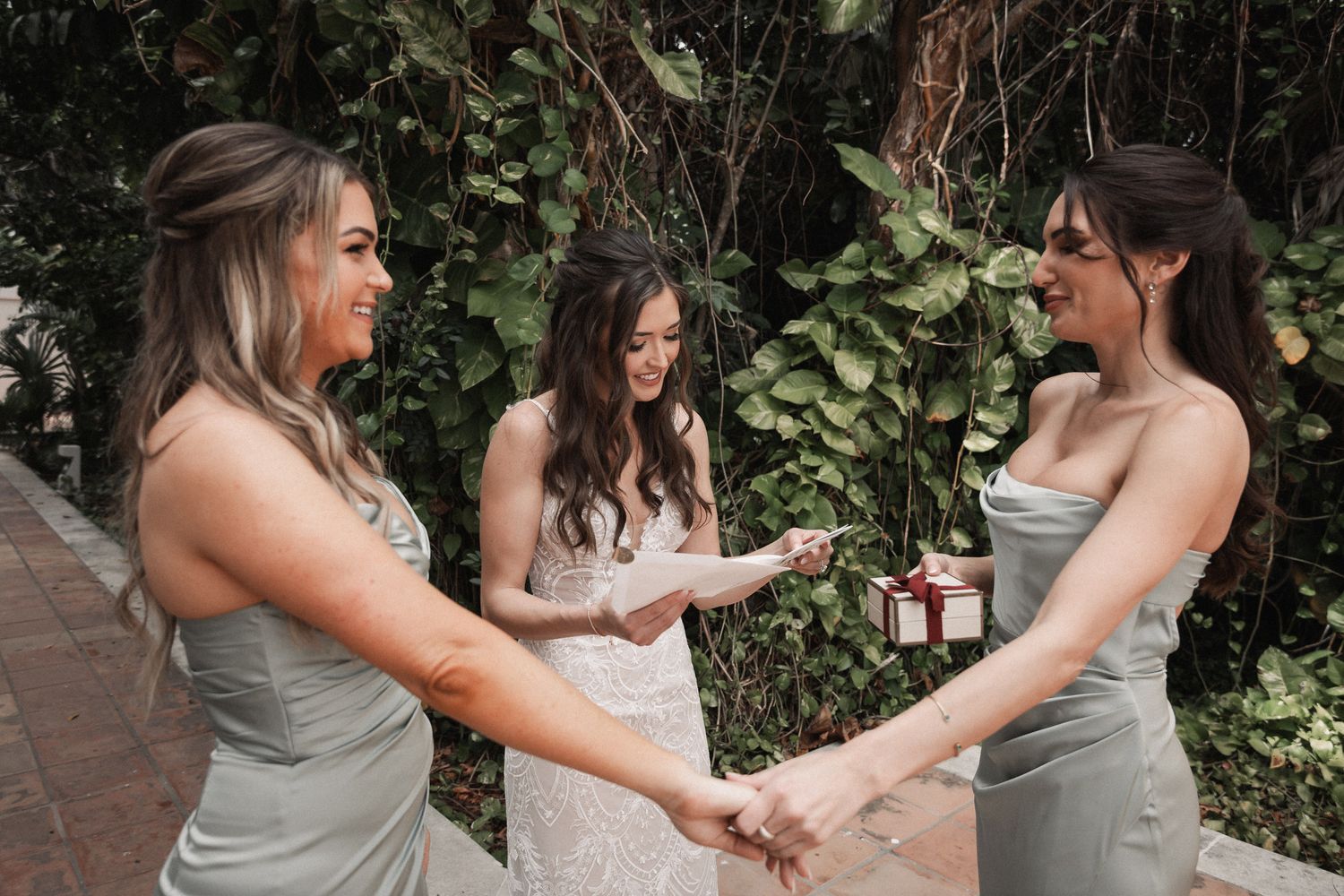 Bride receiving a gift from smiling bridesmaids in sage green dresses amid tropical greenery.