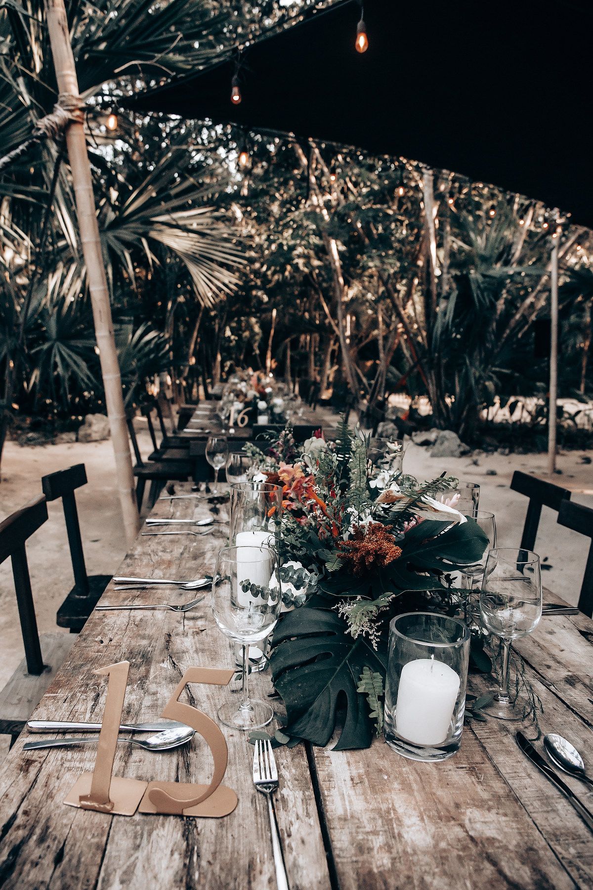 Elegant table setting with dark monstera leaves and wine glasses on a rustic wooden table under string lights.