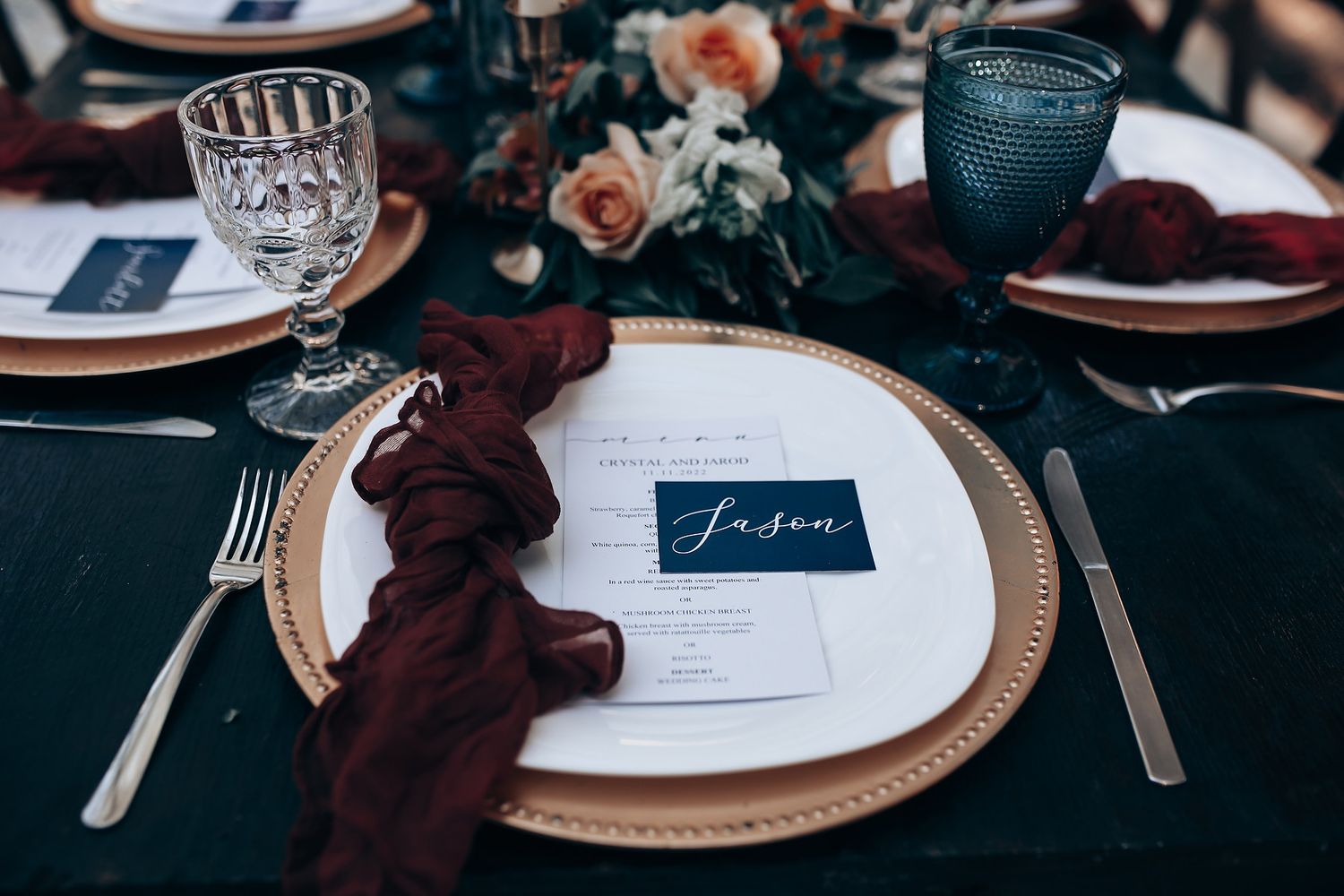 Elegant wedding table setting with burgundy napkin, crystal glasses and place card on gold charger plate.