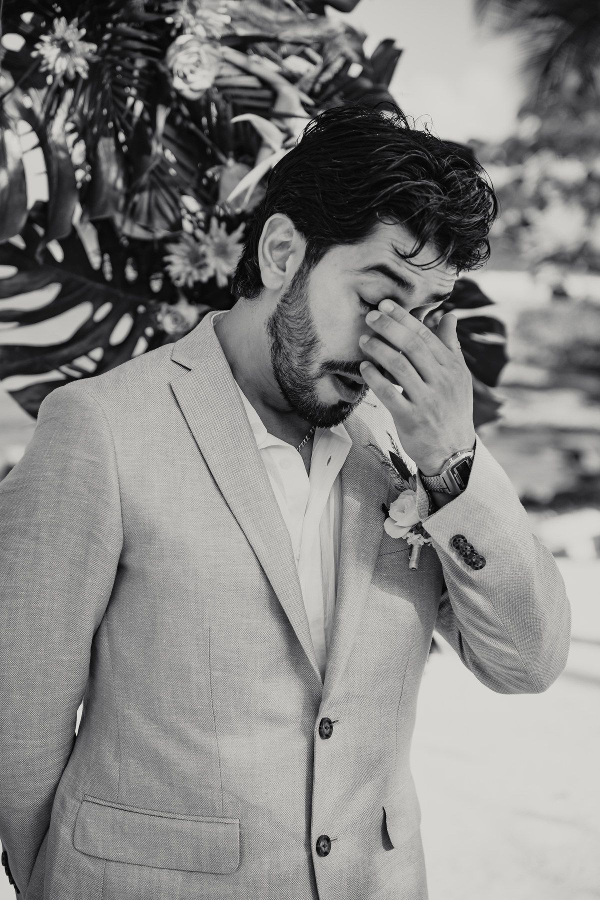 Tearful groom in gray suit covering face with hand at outdoor beach wedding ceremony.