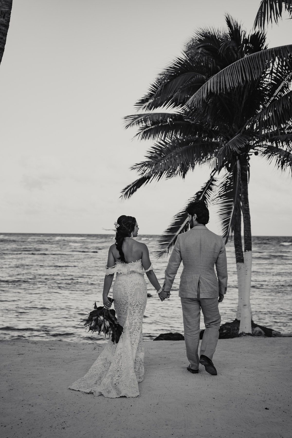 Bride and groom holding hands walking along tropical beach at sunset with palm trees and ocean view.