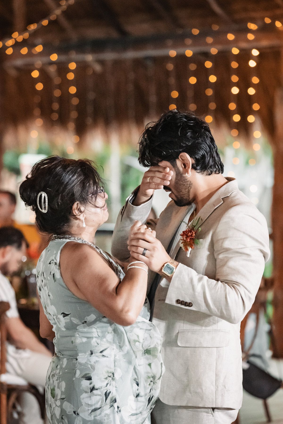 Groom sharing emotional mother-son dance under warm string lights at tropical beach wedding reception.