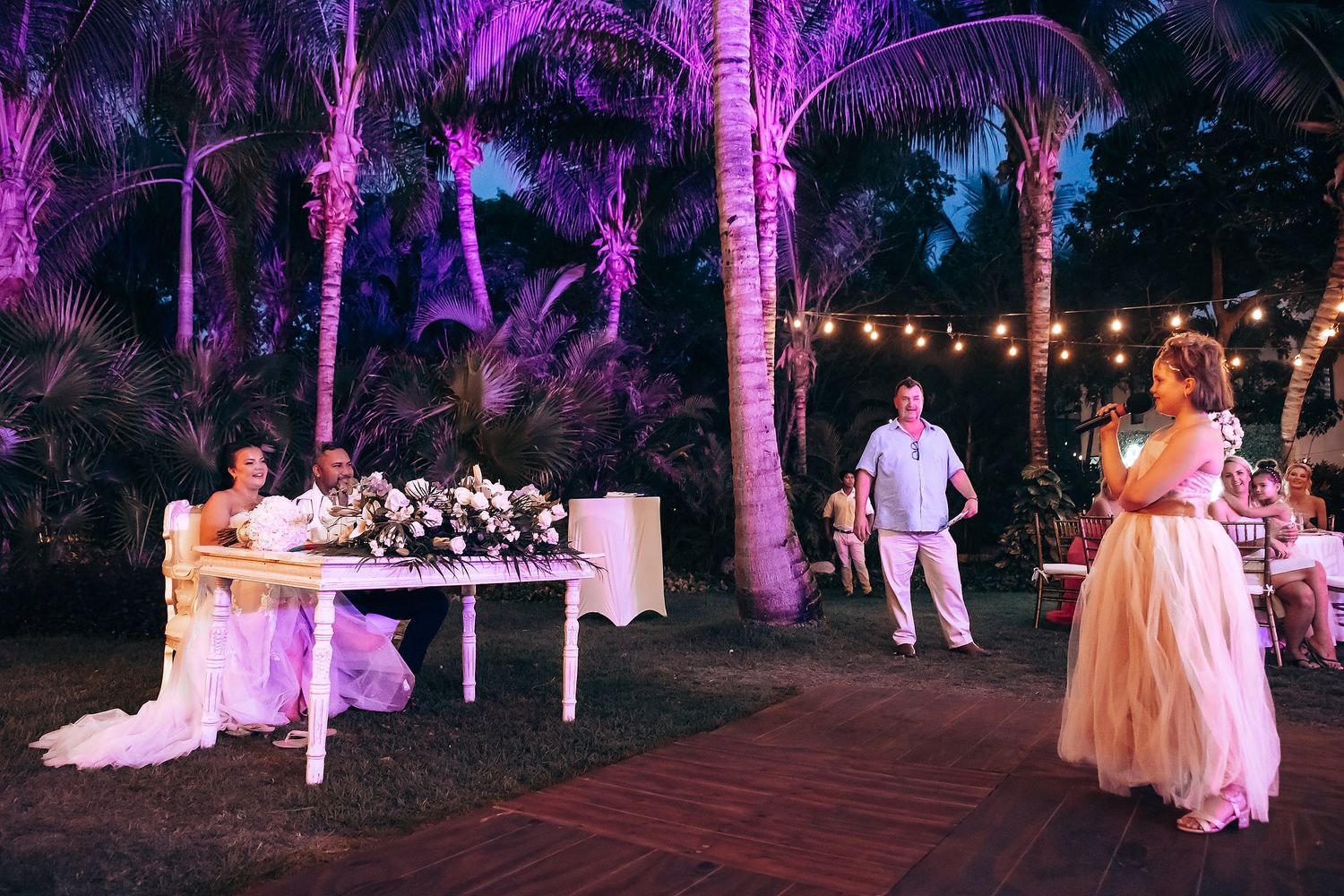Purple-lit palm trees create a magical ambiance at an outdoor evening wedding celebration.