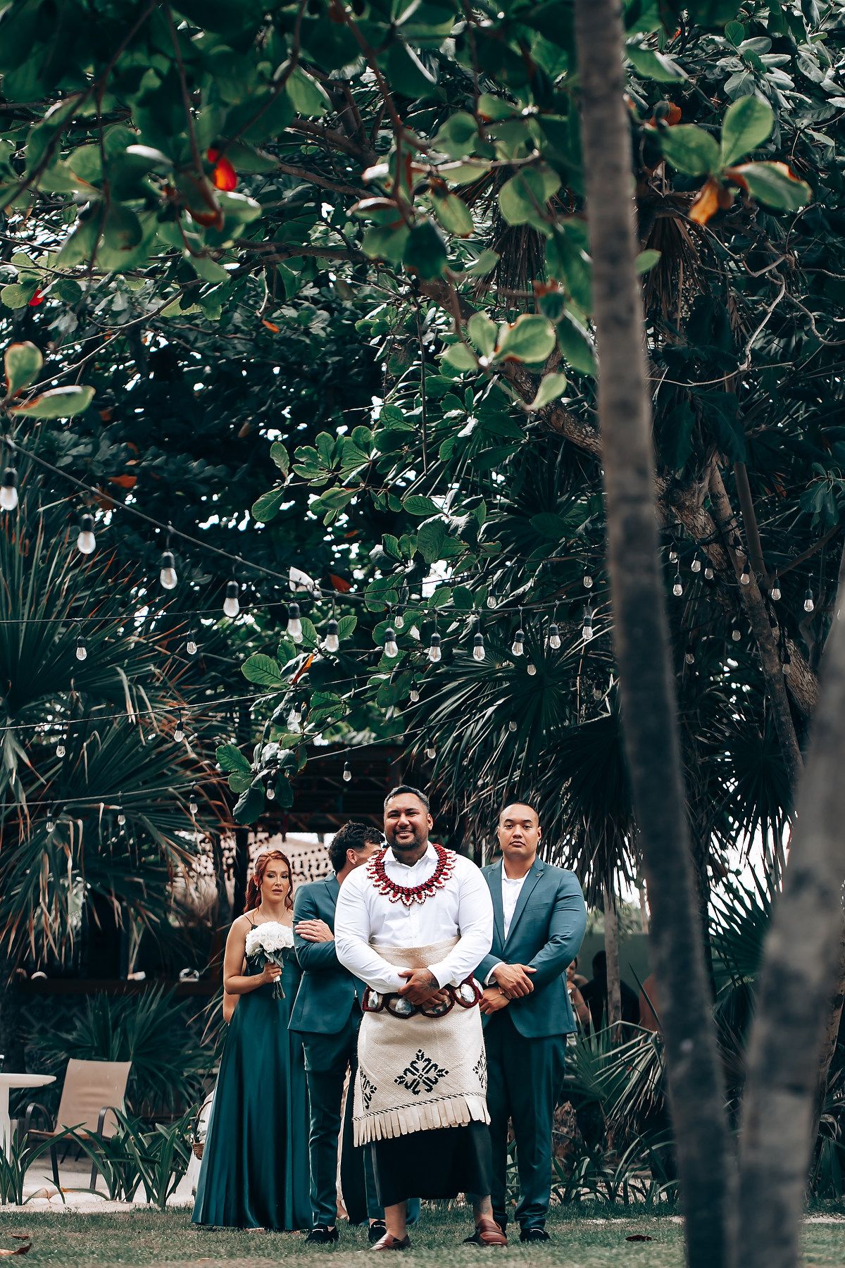 A tropical outdoor wedding ceremony setup with guests seated under palm trees and greenery.