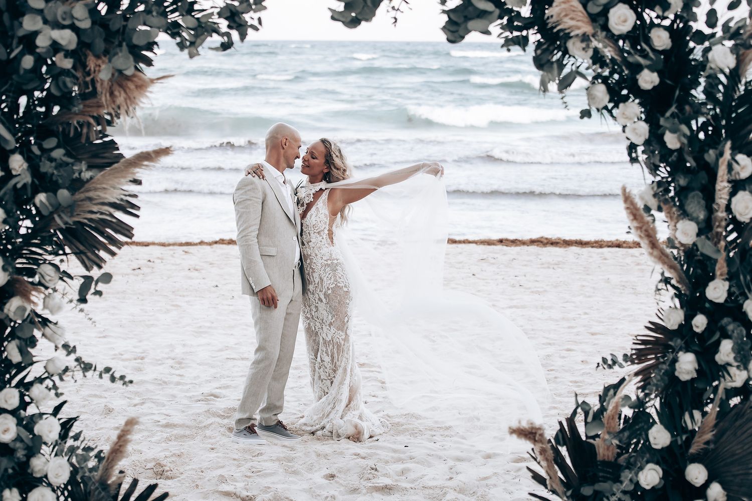 A couple in light-colored attire embrace on a beach under a floral arch during their wedding.