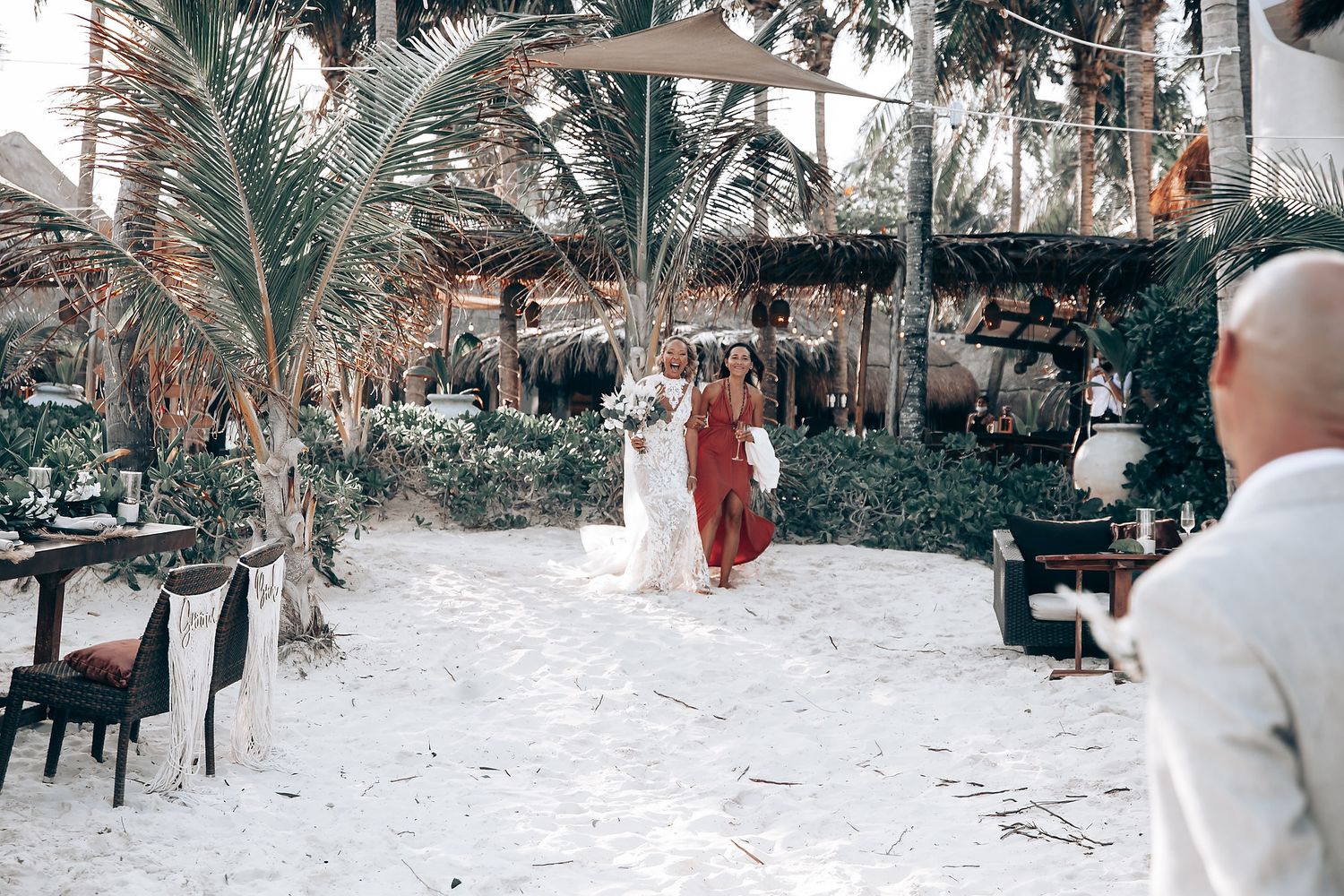 A beach wedding ceremony setup with palm trees and white chairs on sandy ground.