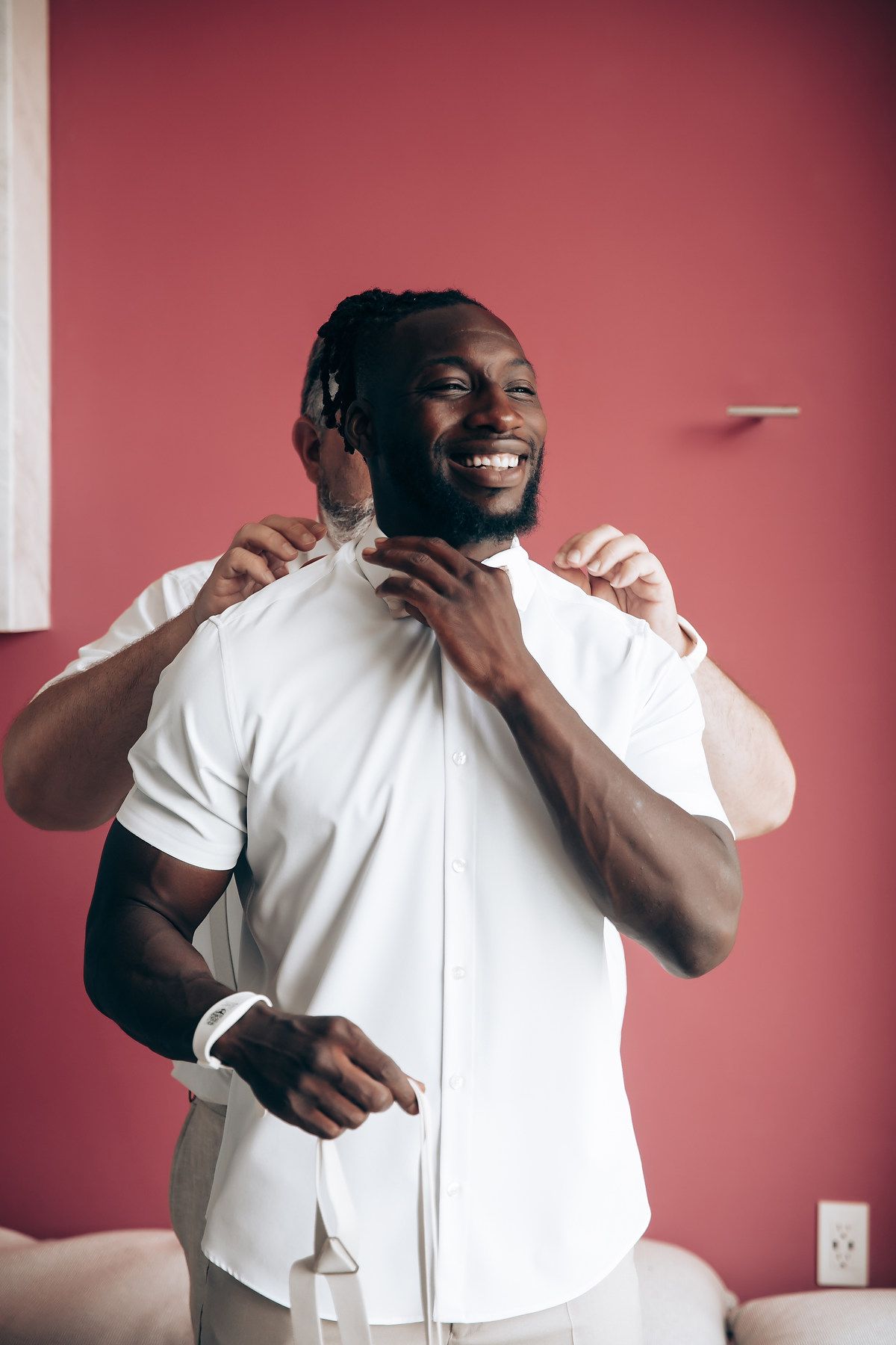 A person in a white button-down shirt adjusts their collar while smiling against a pink wall.