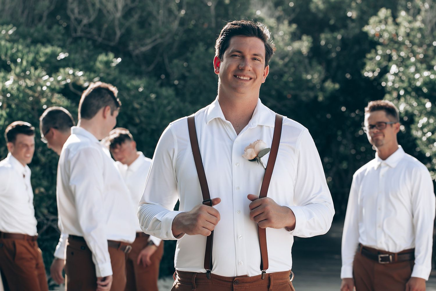 A group in white shirts and brown suspenders stands together outdoors on a sunny day.