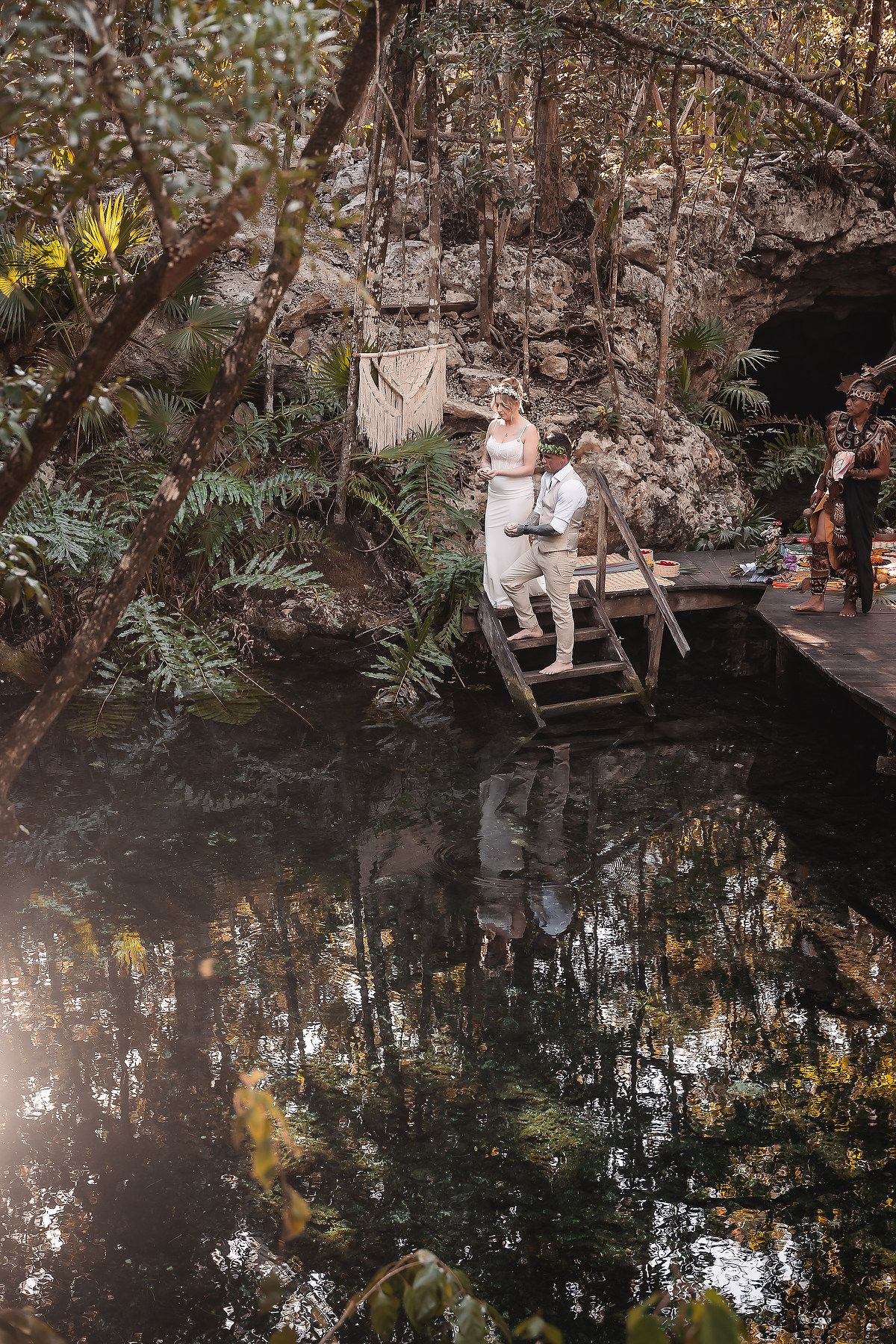 Couple stands on wooden dock by tranquil cenote surrounded by lush jungle foliage during outdoor wedding ceremony.