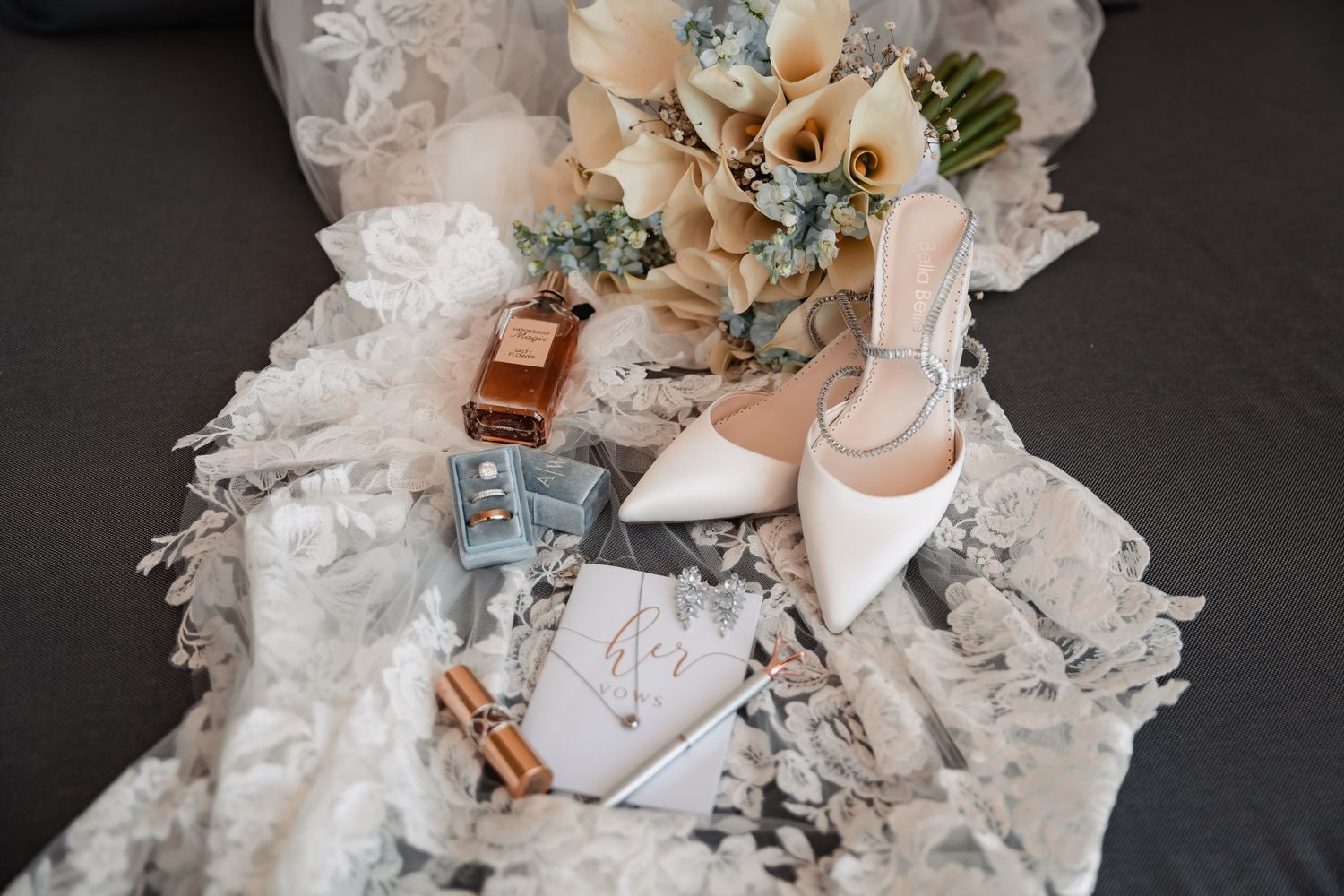 Elegant wedding flat lay with white heels, lace bridal gown, floral bouquet, perfume, and accessories on dark background.