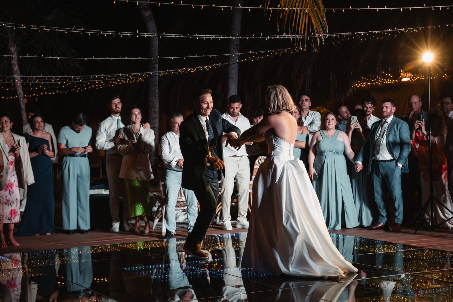 Bride and groom share first dance surrounded by guests in blue attire under string lights at reception.