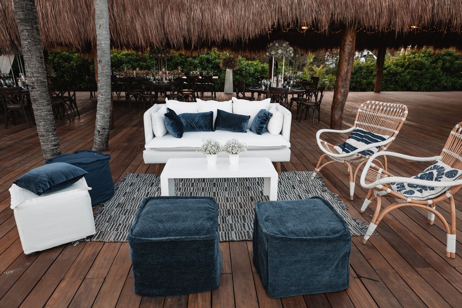 Elegant outdoor wedding lounge area with white sofa, navy ottomans, and rattan chair on wooden deck.