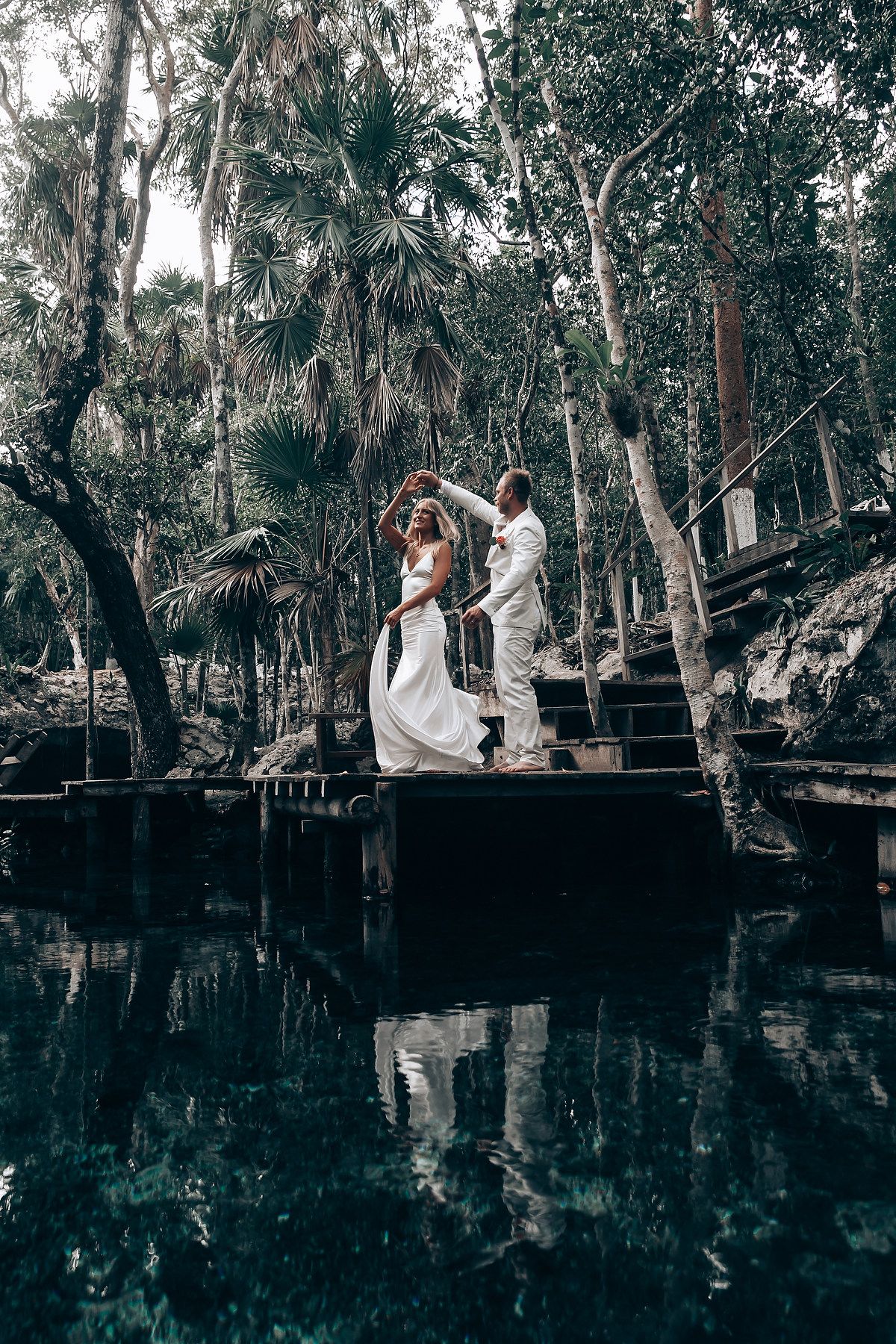 A wedding couple stands on a wooden platform overlooking a serene cenote surrounded by palm trees.