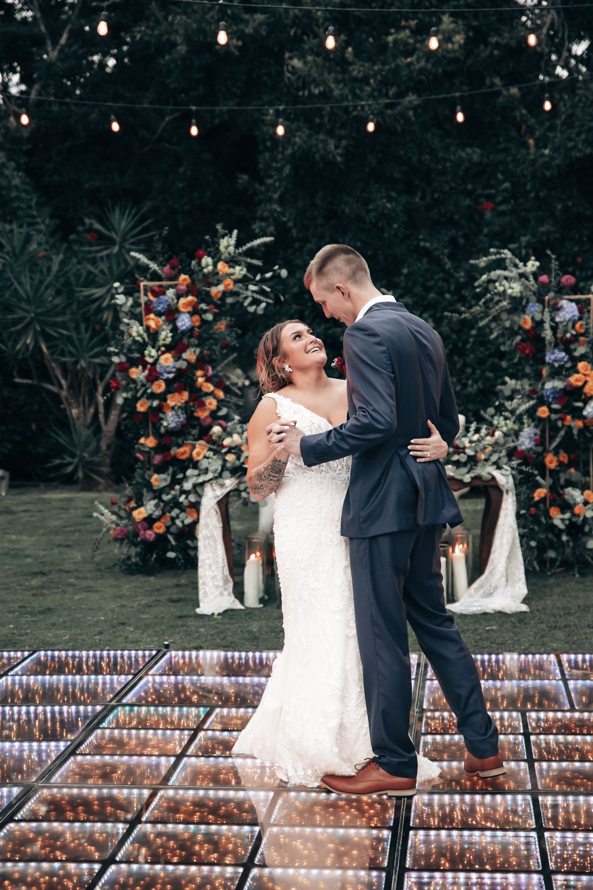 Bride and groom share first dance on mirrored dance floor surrounded by colorful floral arrangements outdoors.
