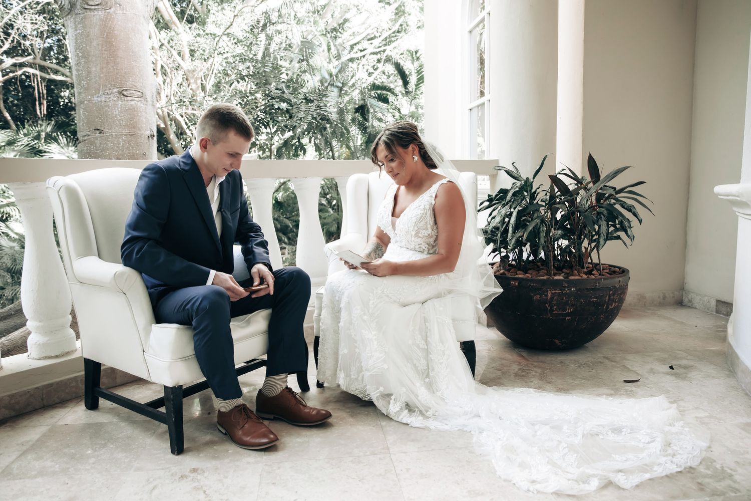 Bride and groom sit in white chairs reading private vows on a sunlit outdoor patio.