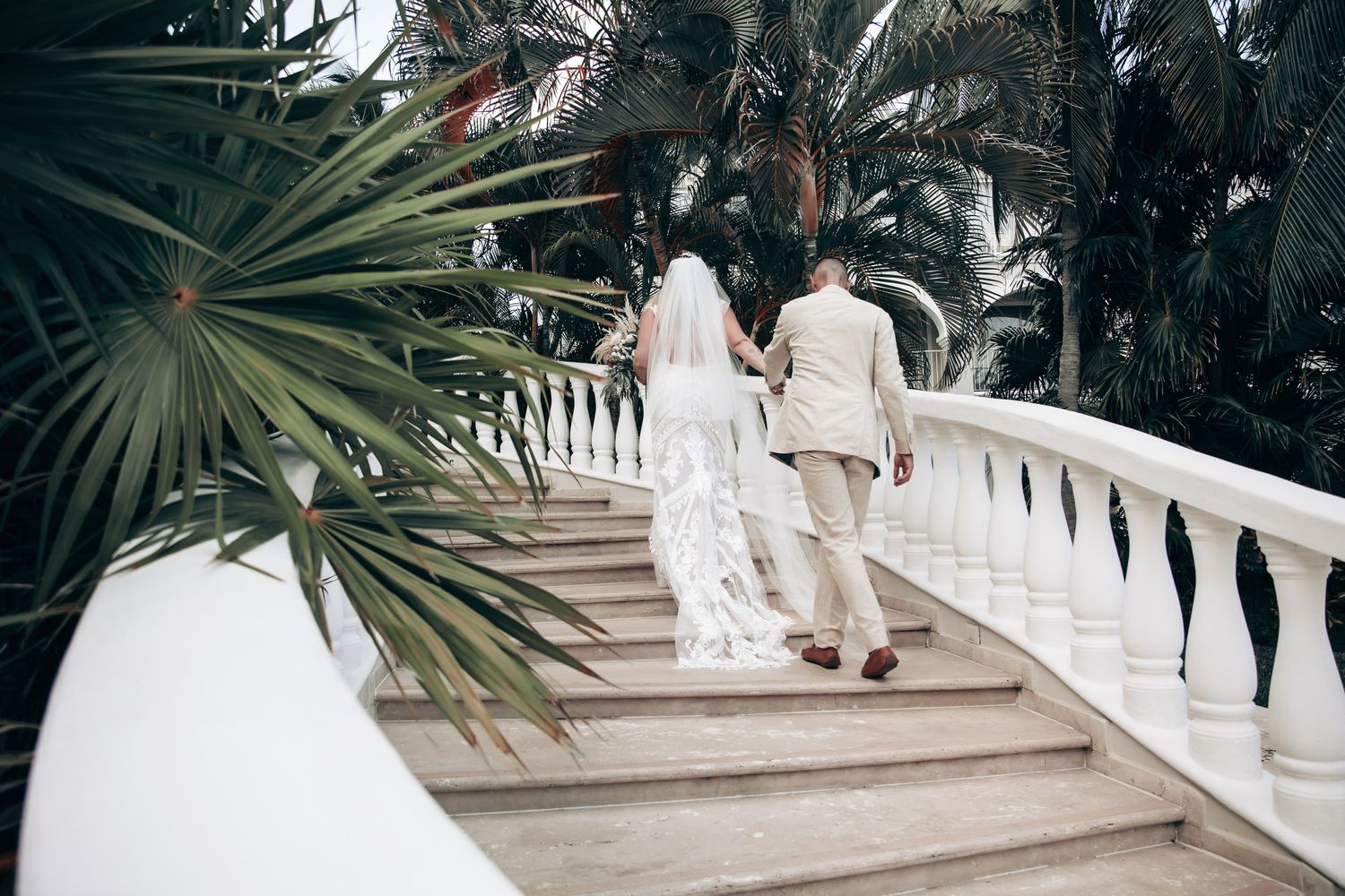 Bride and groom walking up elegant white staircase surrounded by tropical palm trees at outdoor wedding venue at Dreams Tulum