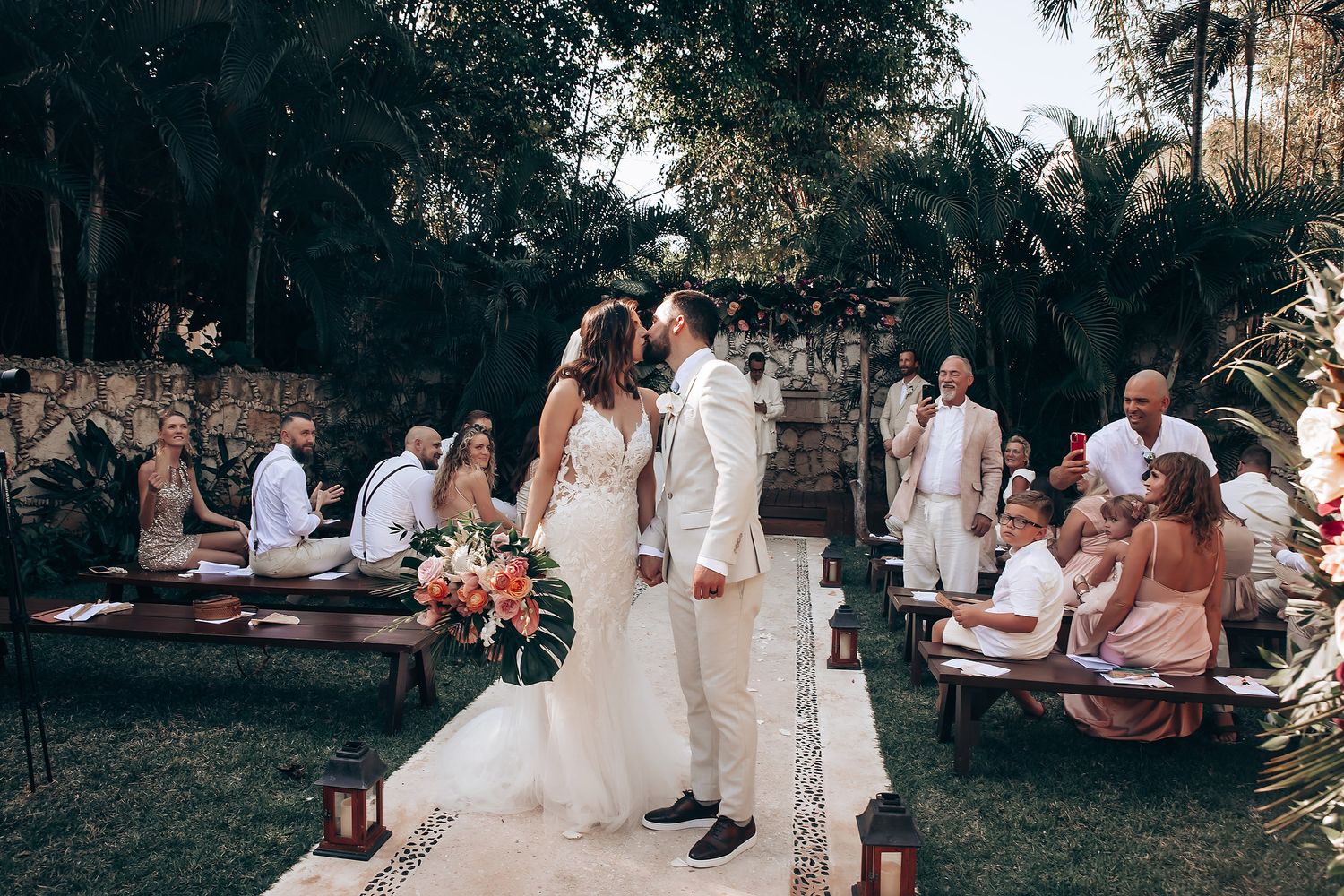 Outdoor wedding celebration with wooden benches and tropical garden backdrop.