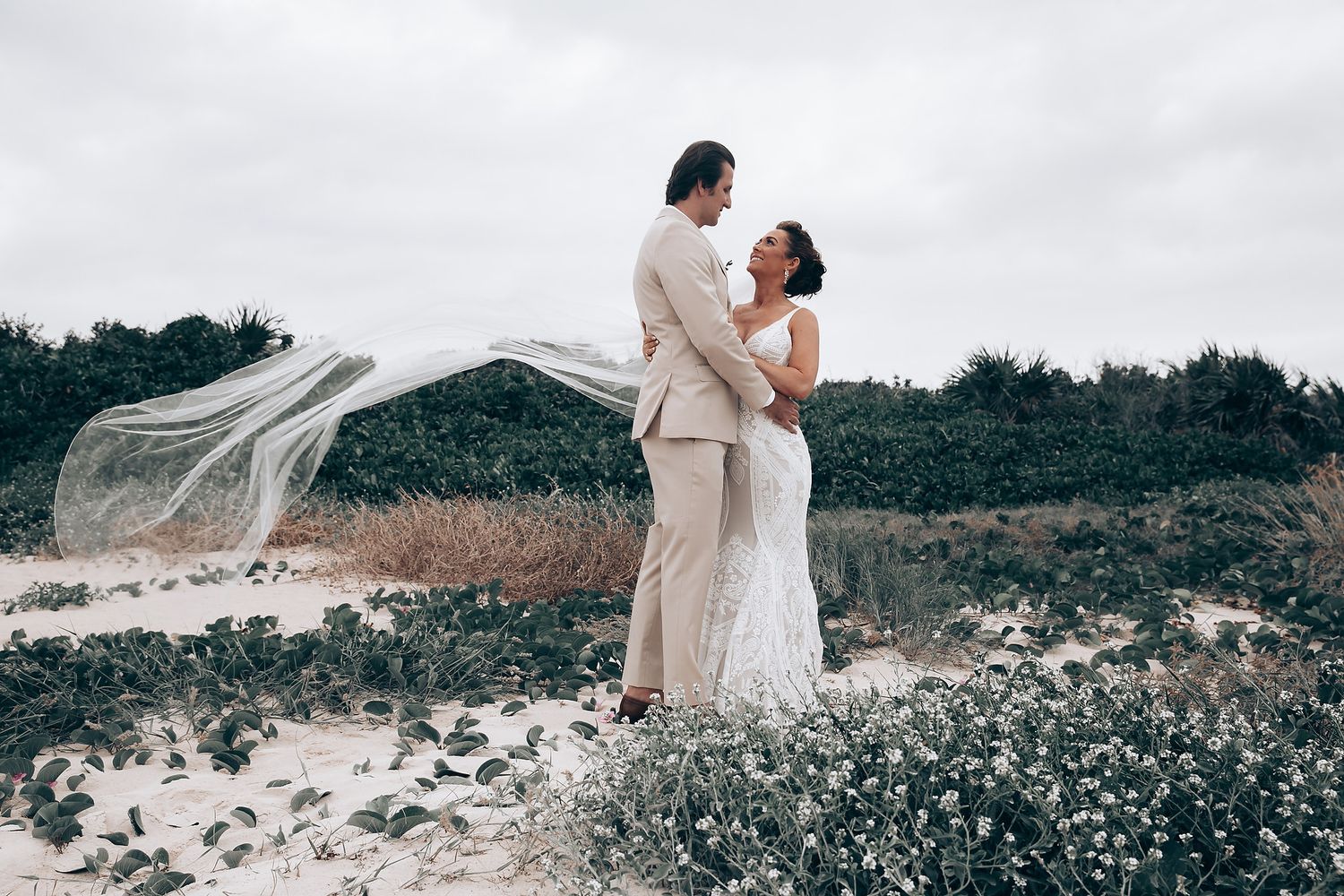 Newlyweds embracing on a scenic hilltop with flowing white dress against cloudy sky.