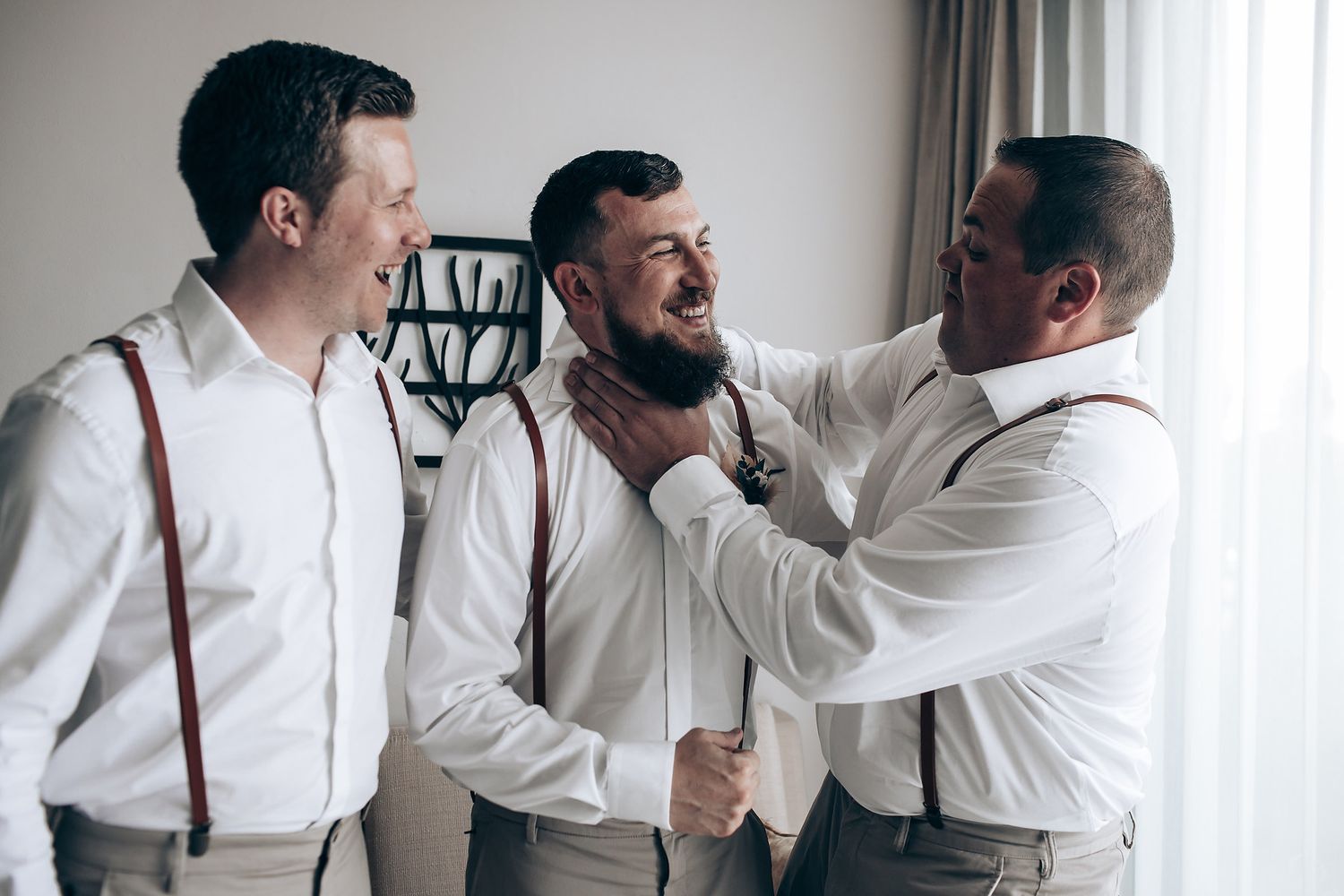 Groomsmen in white shirts and suspenders help each other get ready before a wedding ceremony.
