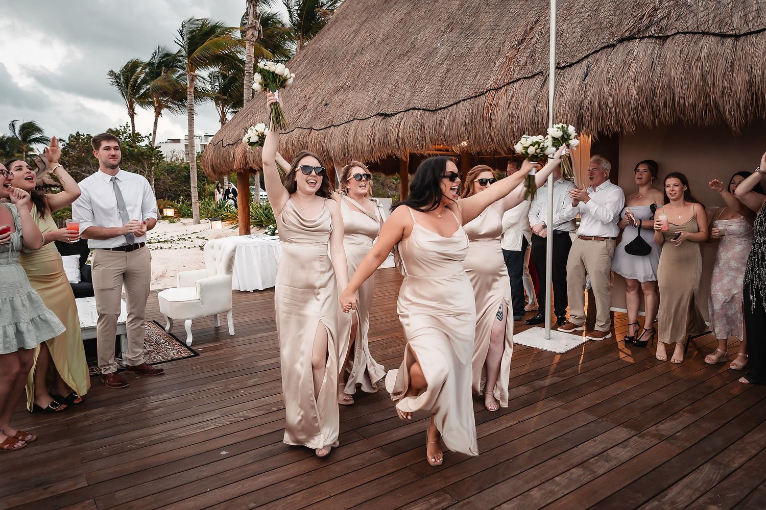 Bridesmaids in champagne gowns dancing and celebrating on tropical resort deck at outdoor beach wedding.
