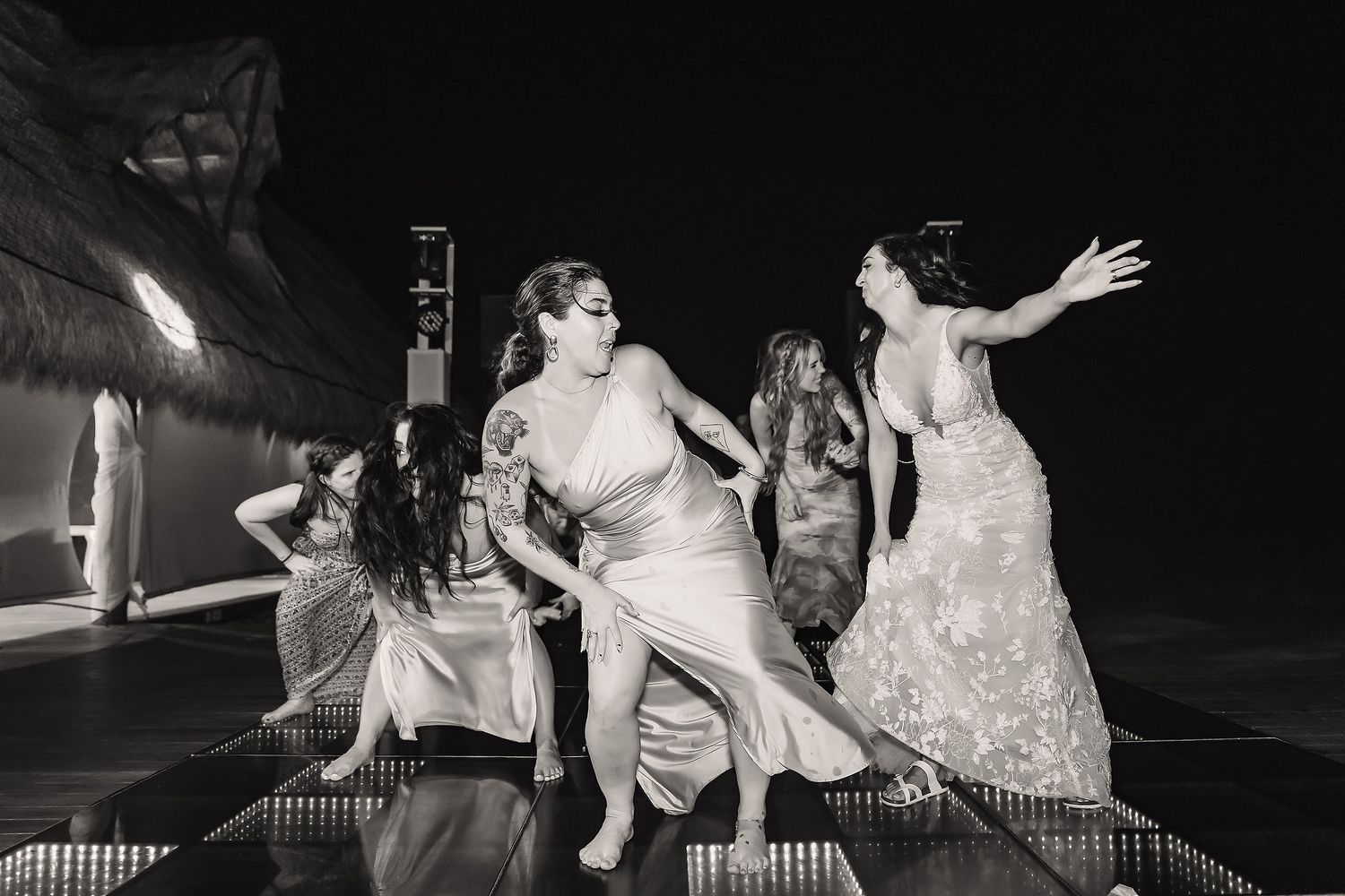 Women in elegant gowns dancing joyfully at an outdoor wedding reception in black and white photography.