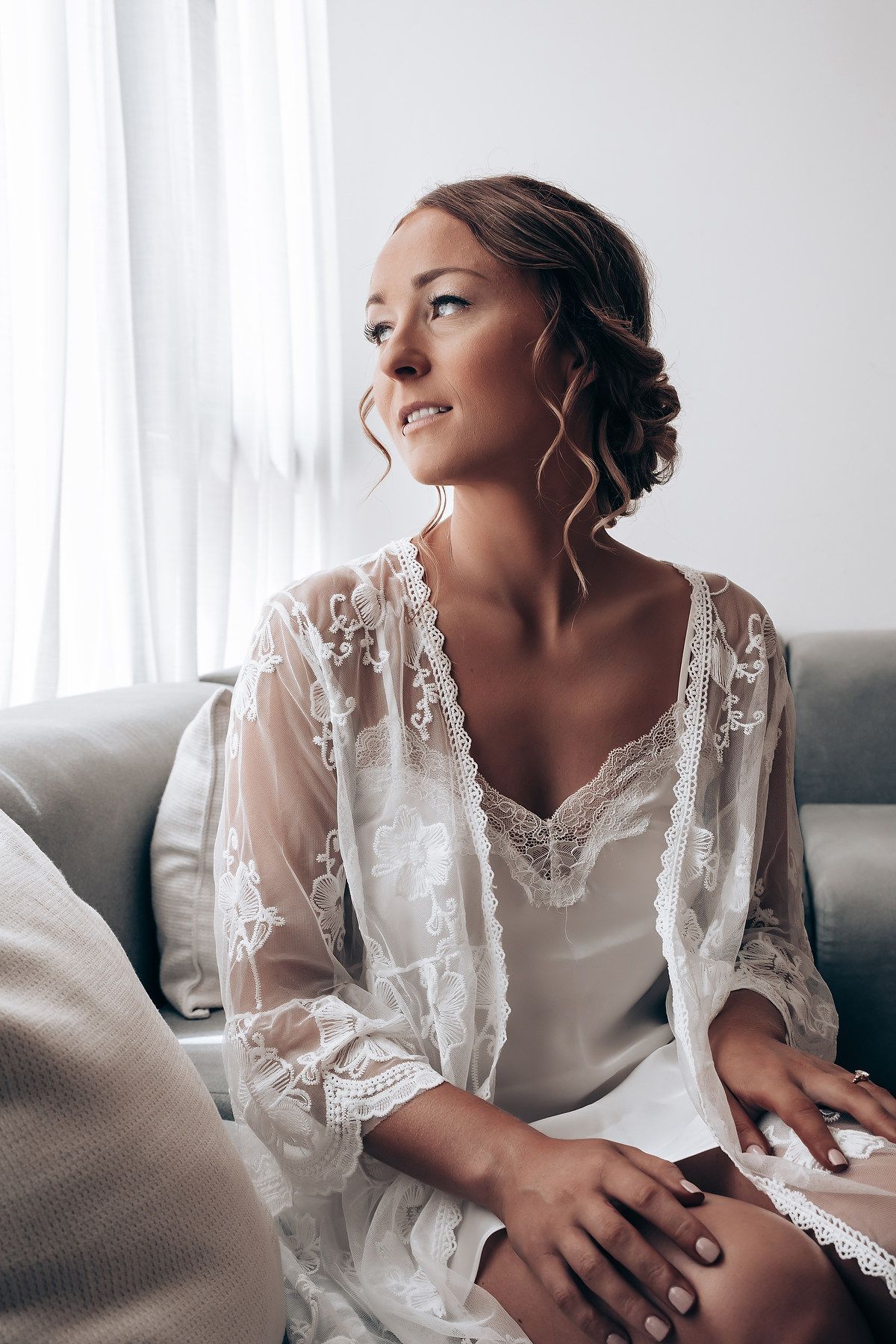 Bride wearing white lace robe relaxing on grey couch in morning light.