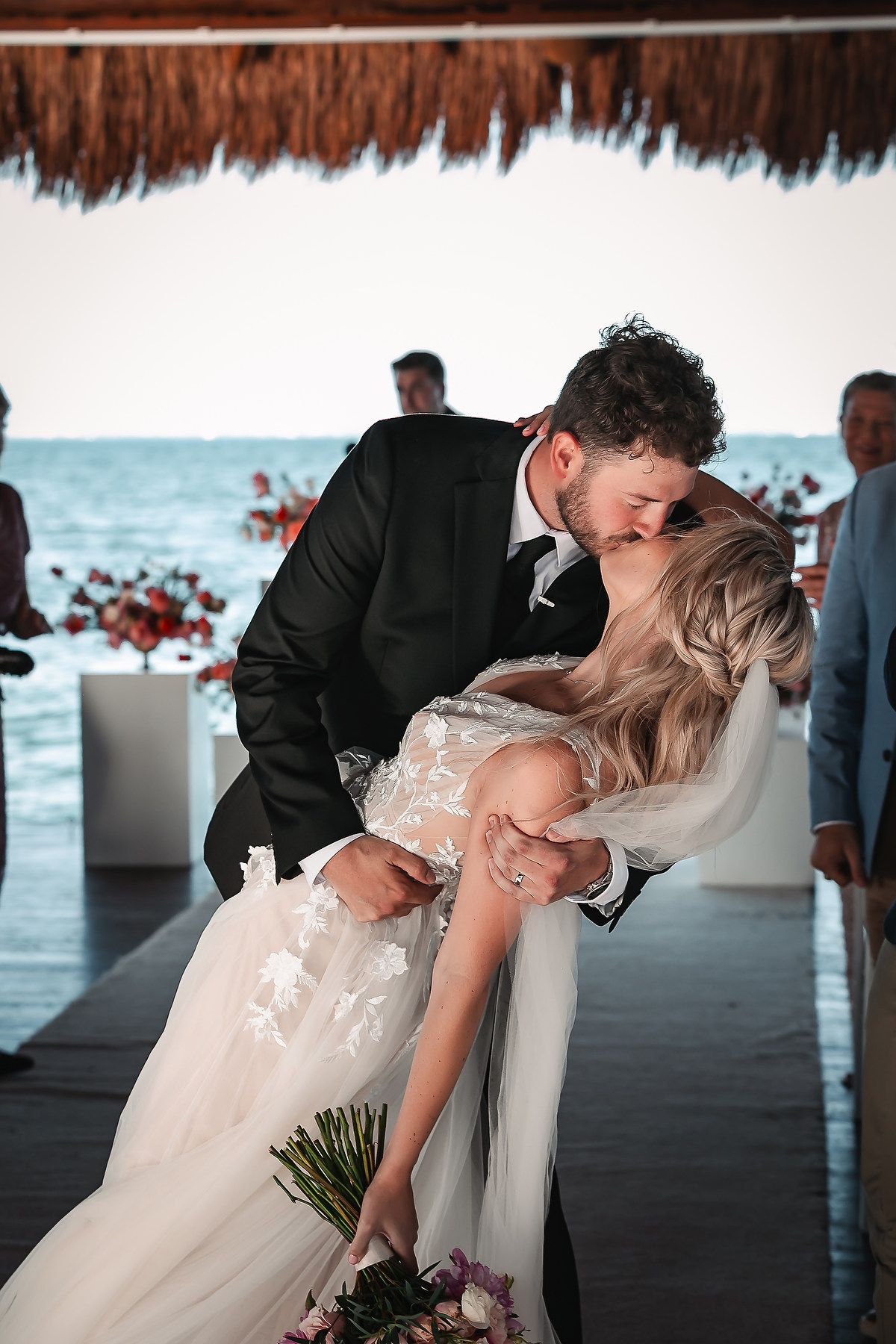 Romantic beach wedding scene with a couple sharing a dip kiss under a thatched roof.