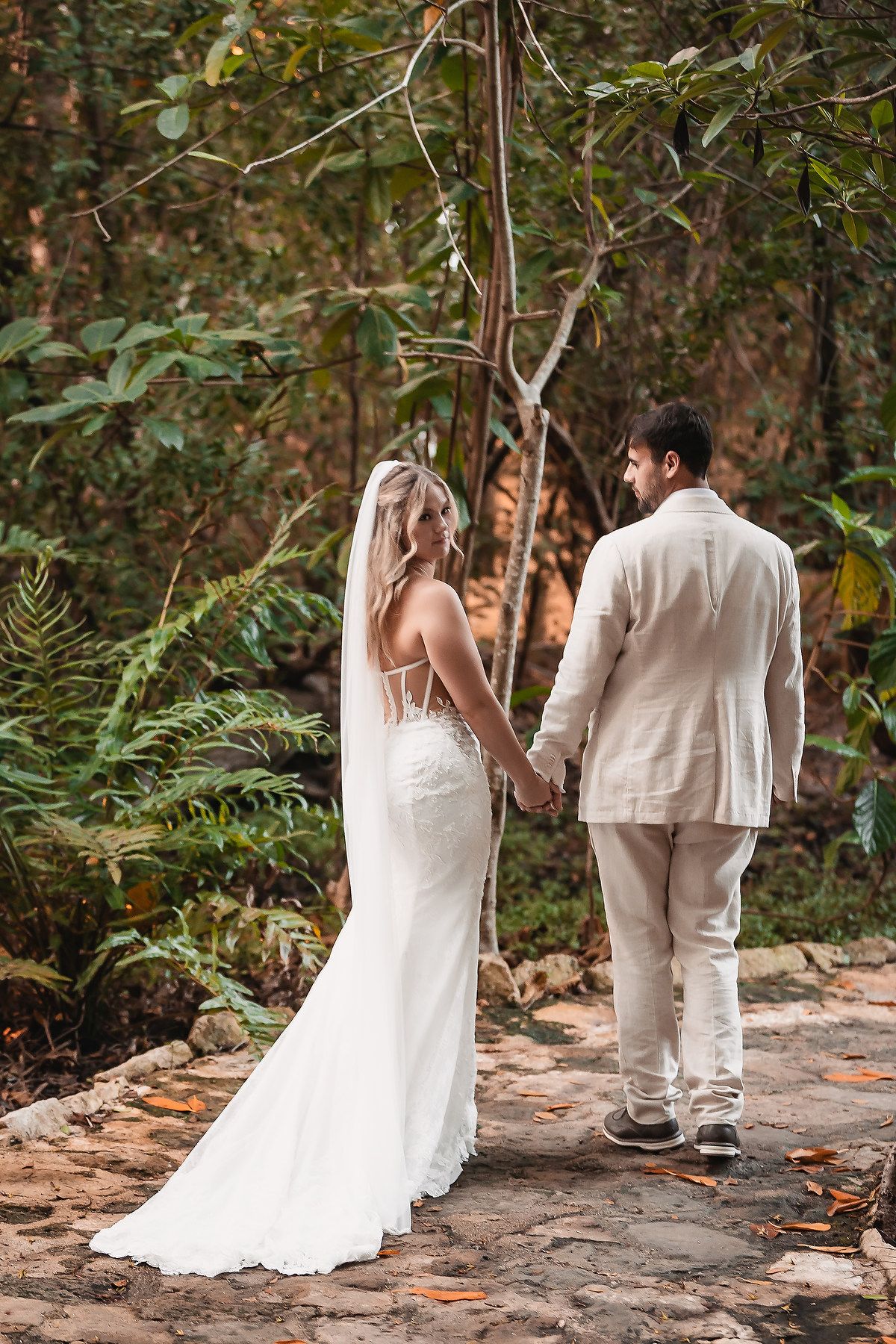 Bride in white lace gown and veil holds hands with groom in cream suit, standing on stone path in lush tropical forest.