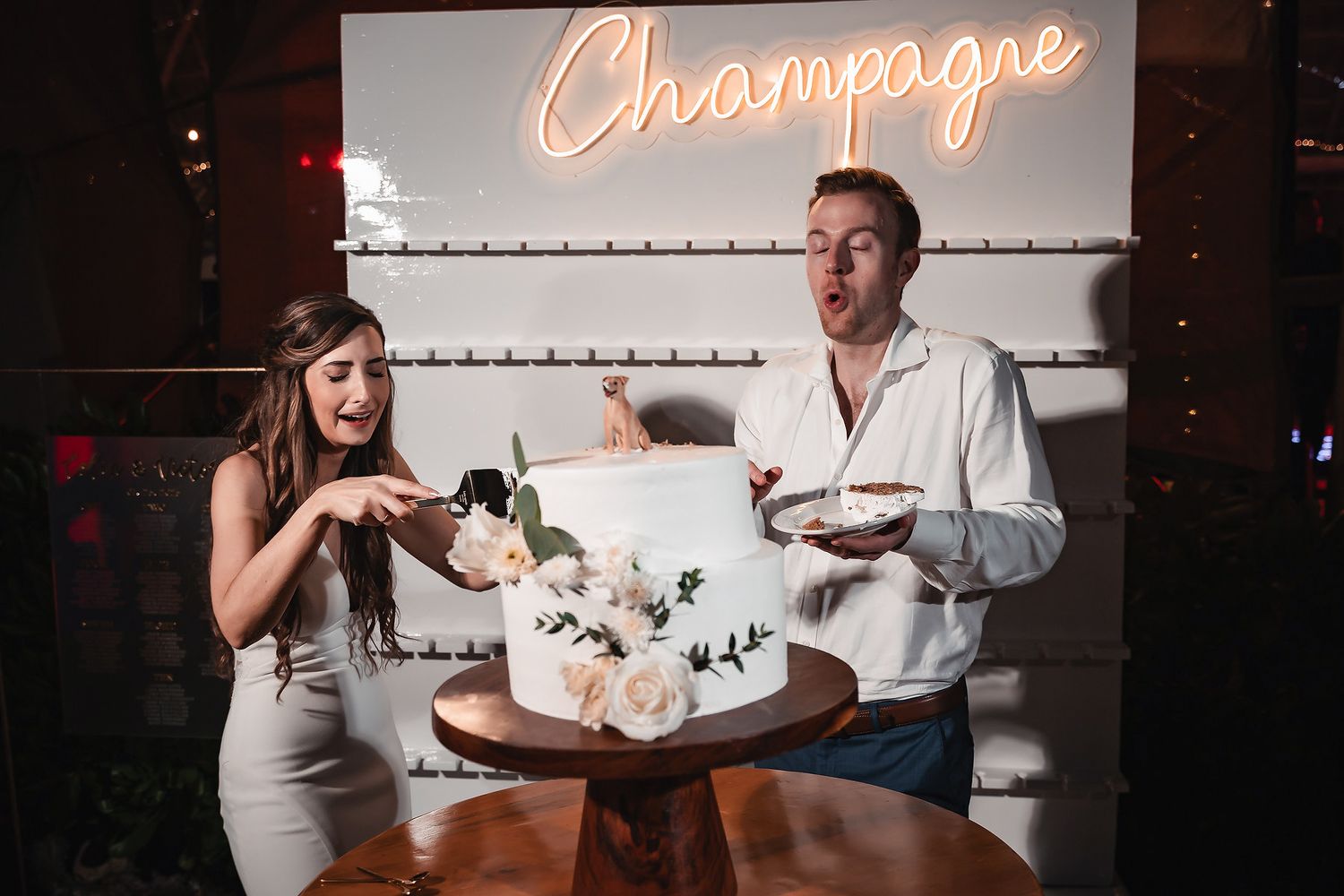 Couple laughing while cutting white floral wedding cake in front of a Champagne neon sign.