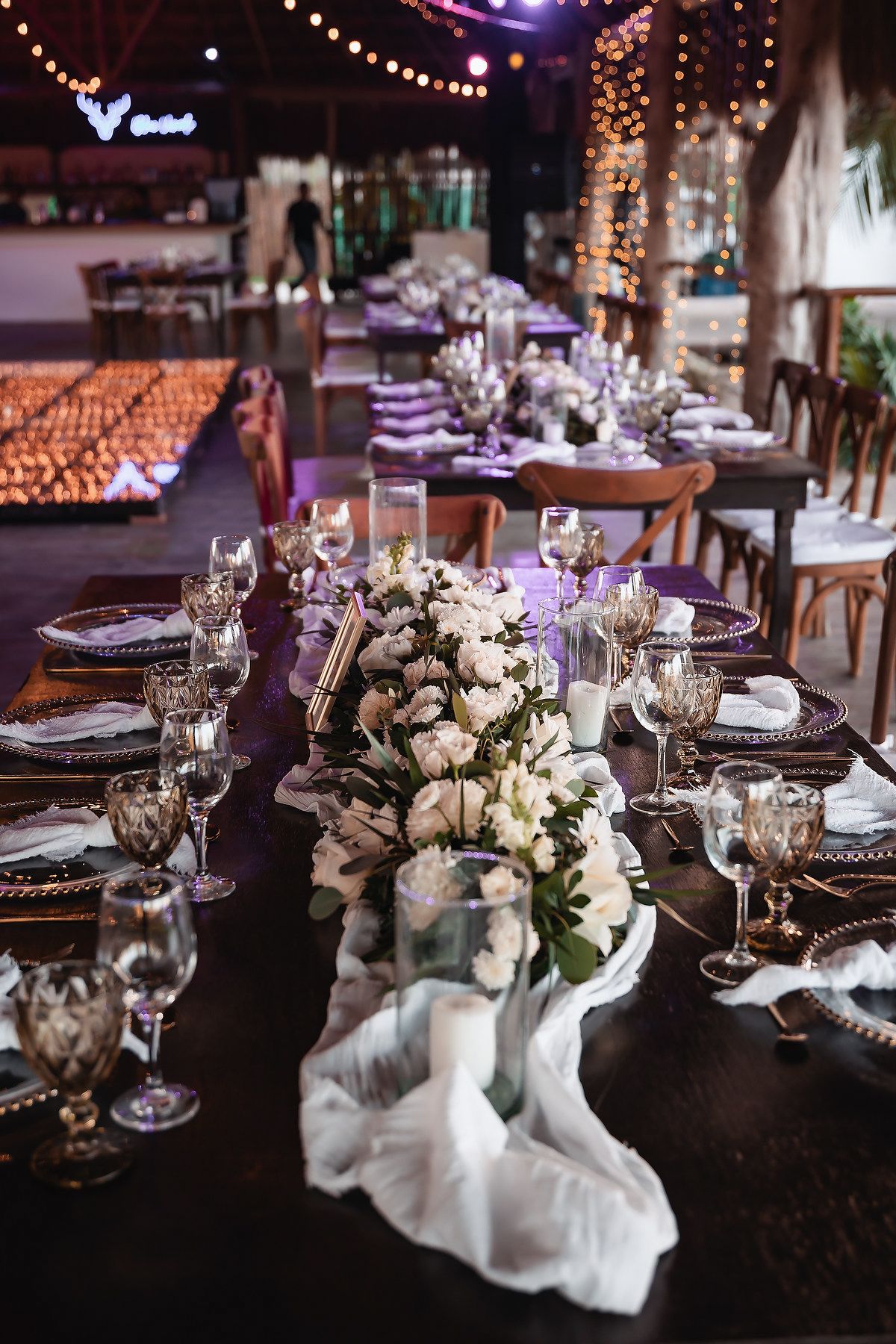 Elegant wedding reception table with white floral centerpiece, crystal glassware, and warm string lights in a decorated venue.