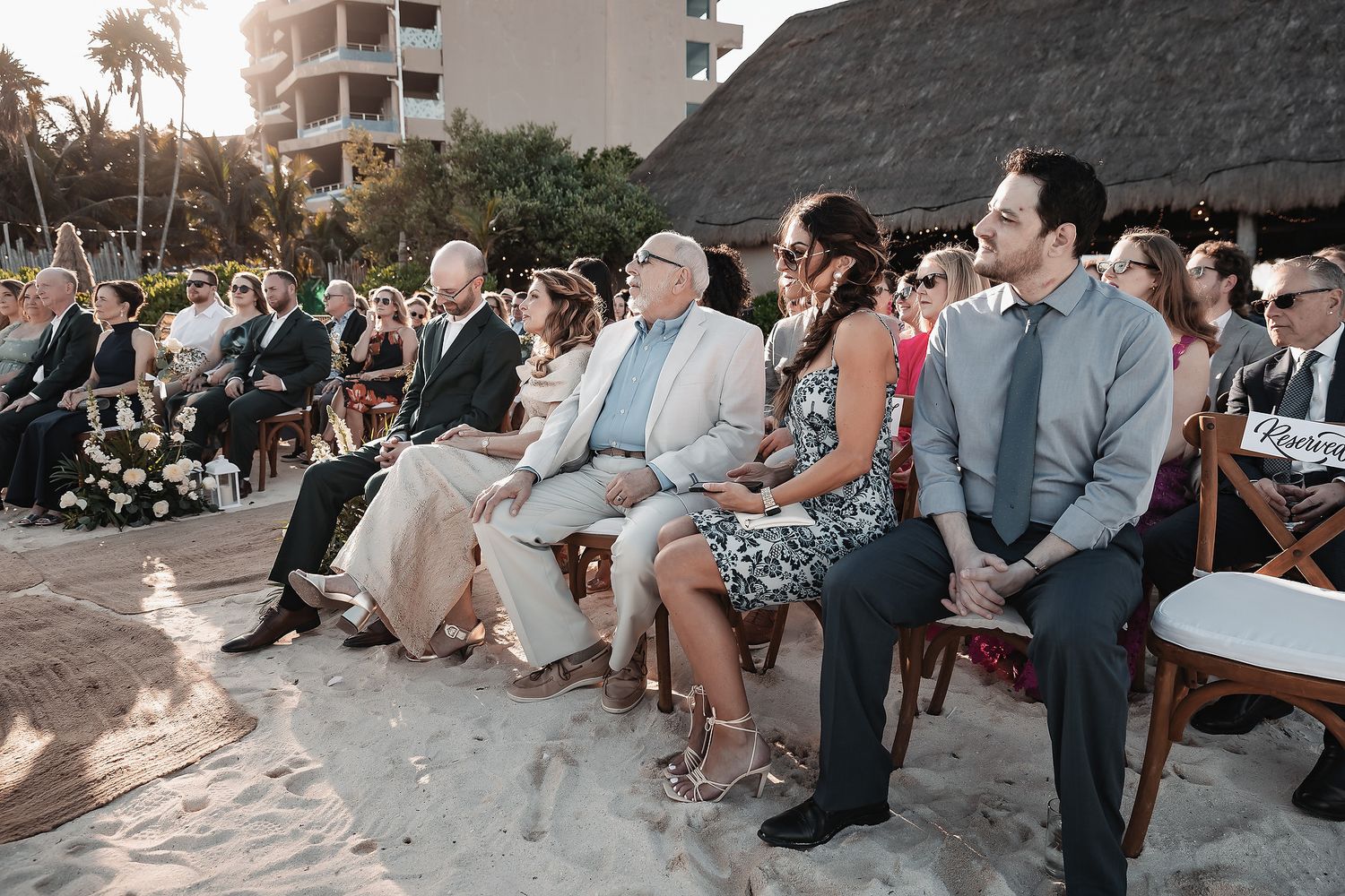 Guests seated on wooden chairs in the sand during an outdoor beach wedding ceremony at a tropical resort.